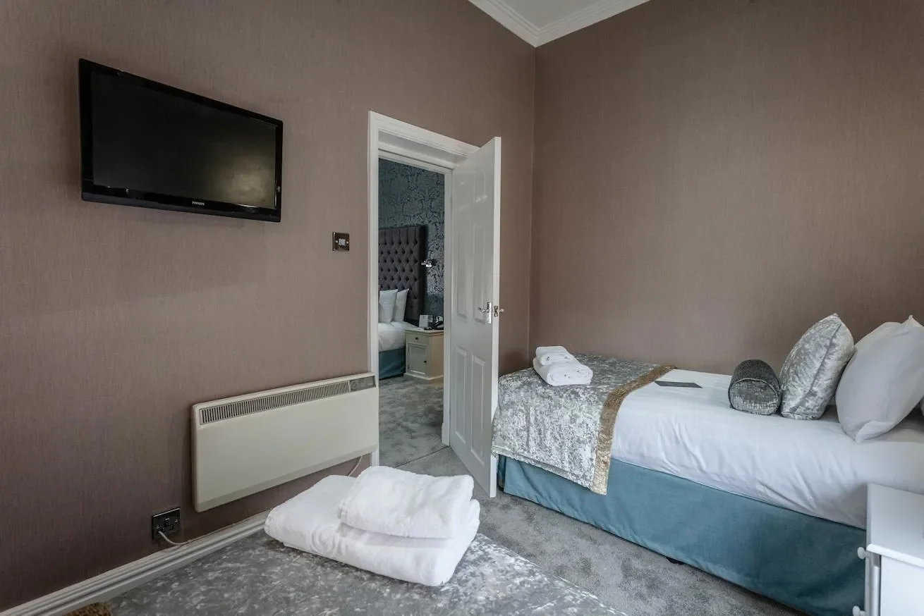 A hotel room with two single beds, pillows, a TV on the wall, a stack of towels on one bed, and a doorway leading to another bedroom with a tufted headboard.