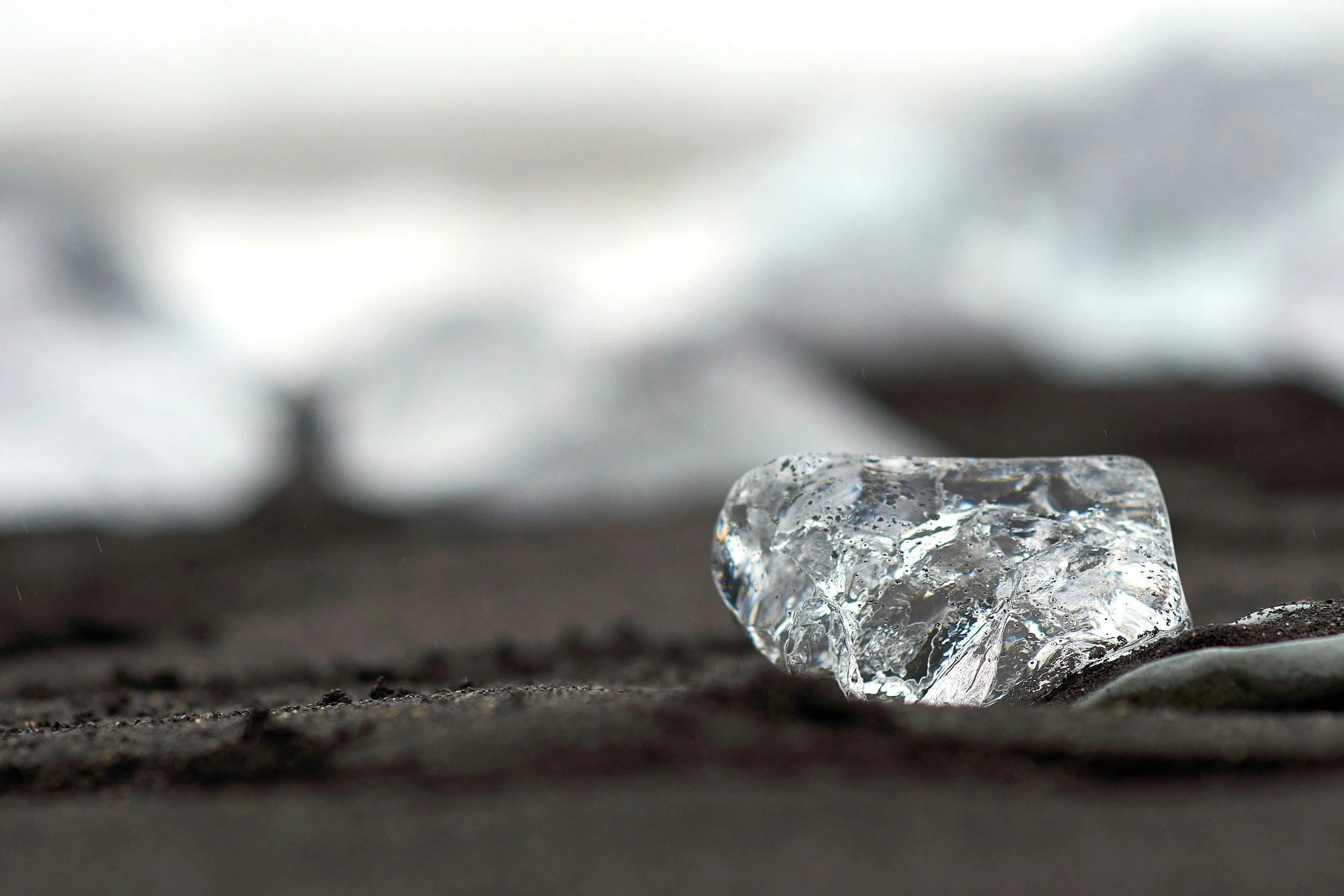 Close-up of a transparent ice cube on dark sand with a blurred background.