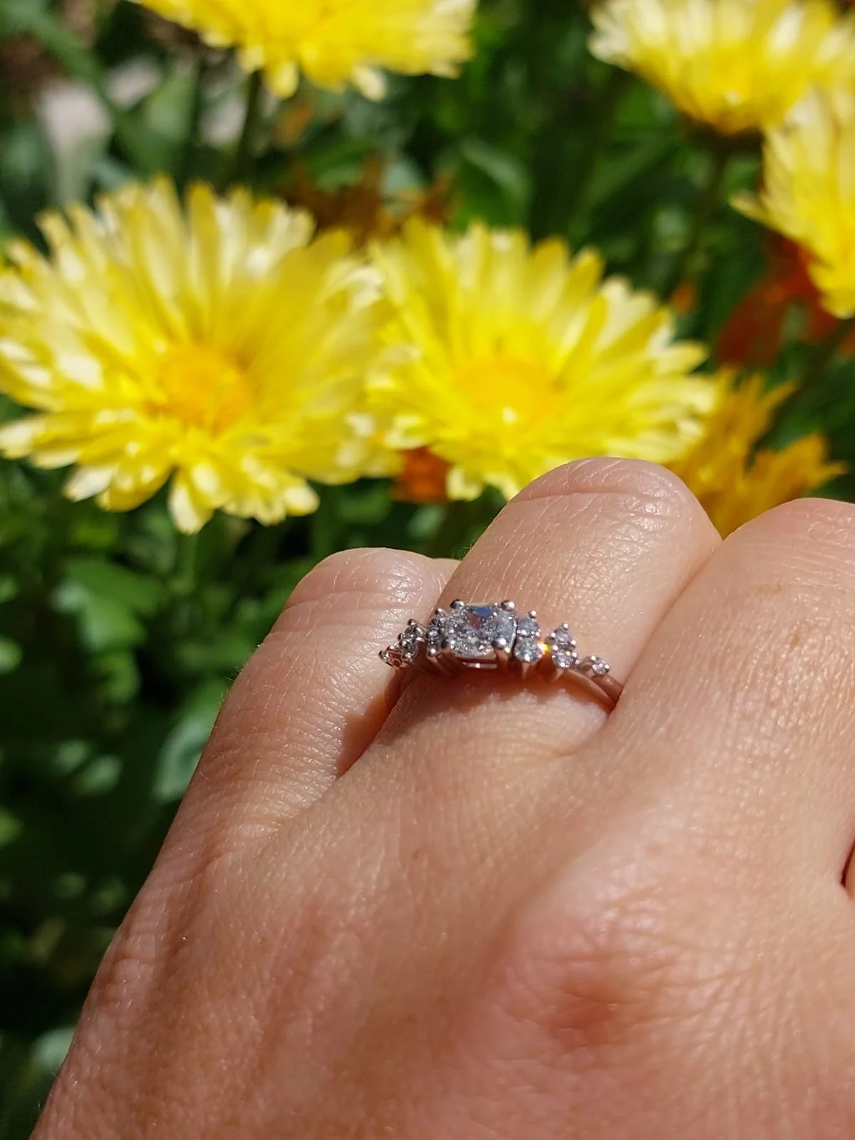 A hand wearing a diamond engagement ring with a yellow flower and green foliage in the background.