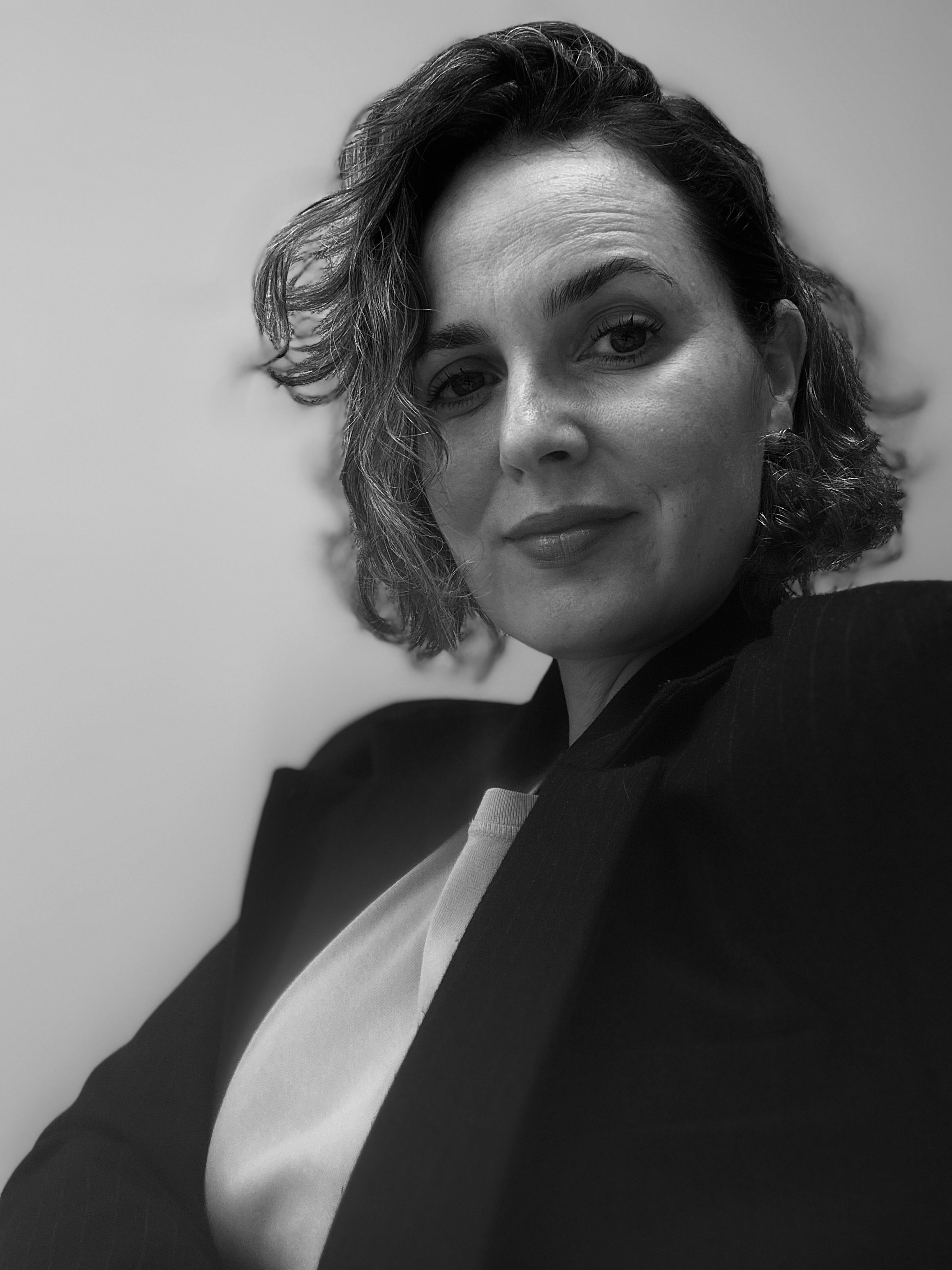 Black and white portrait of a woman with wavy hair, wearing a blazer and a light-colored top, looking slightly to the side with a confident expression.
