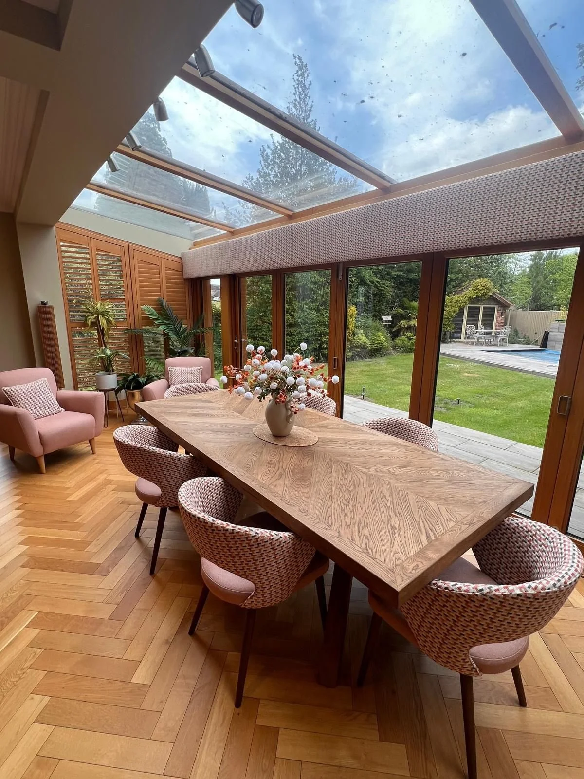 Dining room with a large wooden table, pink chairs around it, a vase with flowers on the table, wood floors, glass sliding doors, and a view of a backyard with a lawn, patio, and pool.