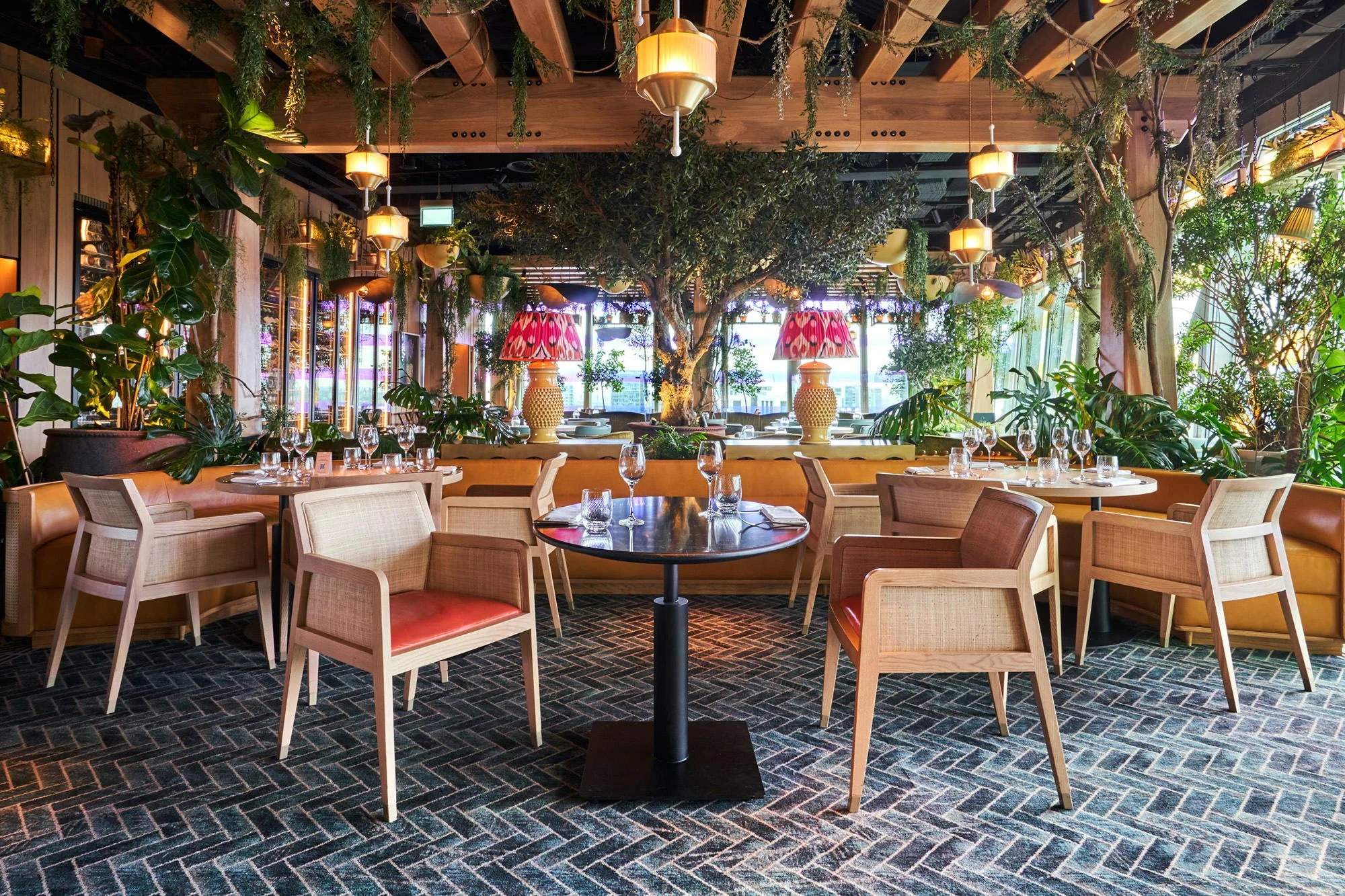 A restaurant with a large central tree, surrounded by tables set with glassware, and plants; decorated with hanging lamps and patterned lamps on windows.