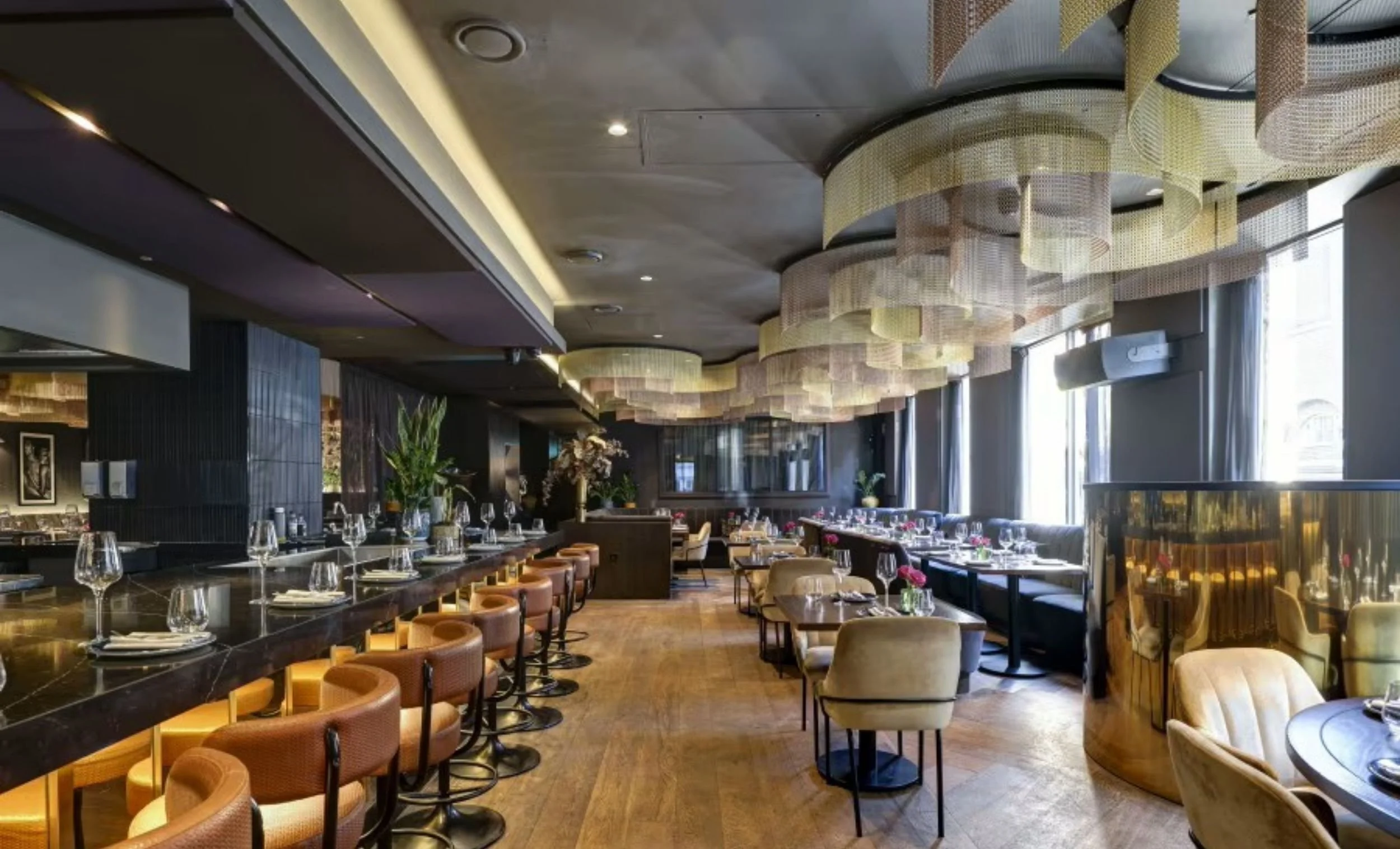Elegant restaurant with a bar counter on the left, tables set with wine glasses and cutlery, large windows on the right, and artistic ceilings with hanging decor.