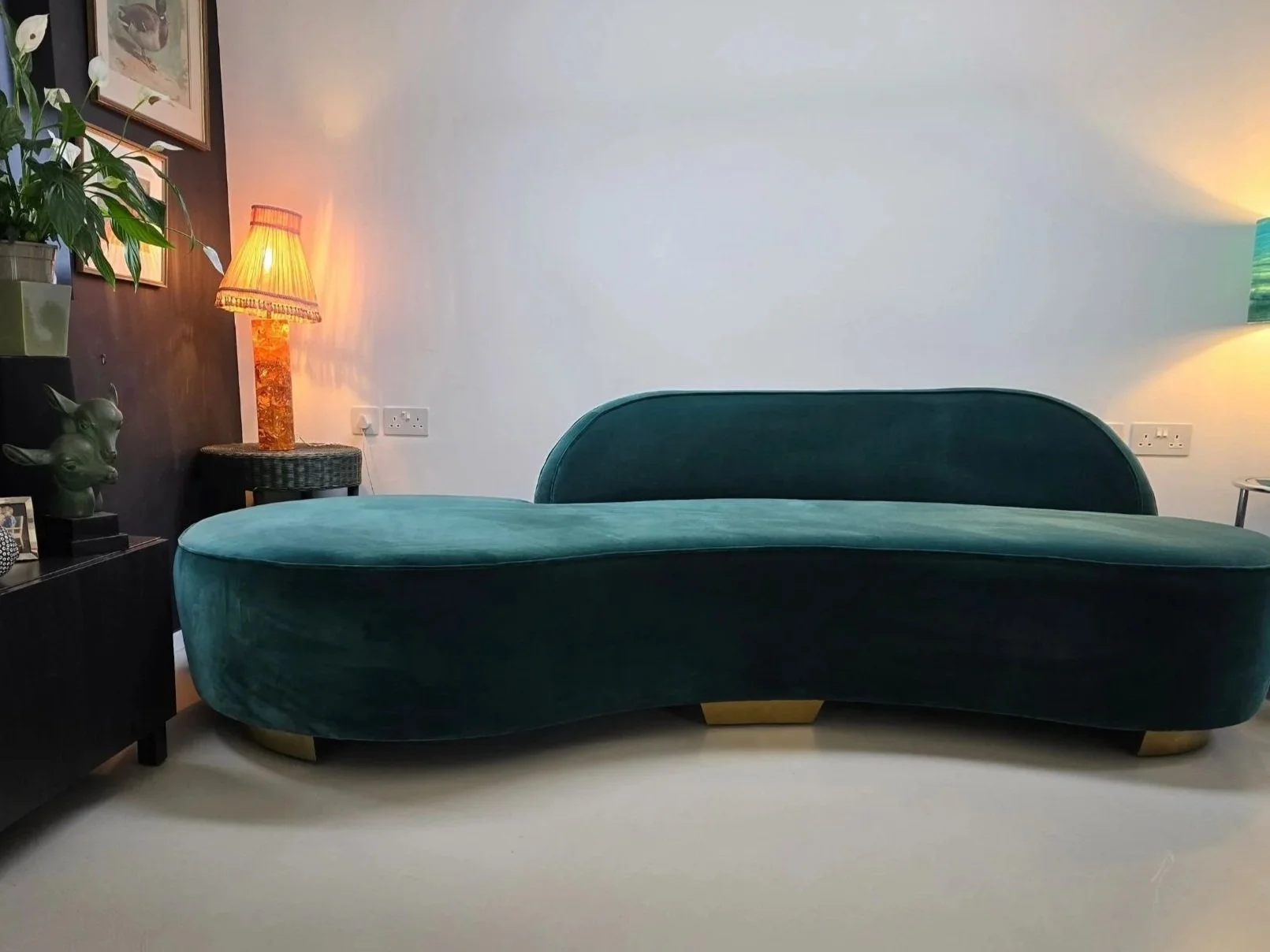 A teal velvet curved sofa in a living room with a dark wood side table, decorative items, and a person on the left with a green ceramic bust of a dog's head. There are two wall-mounted lamps, one with a orange shade and the other with a multicolored shade. The background features a white wall and a dark wood panel with framed pictures, and a potted plant on top of the side table.