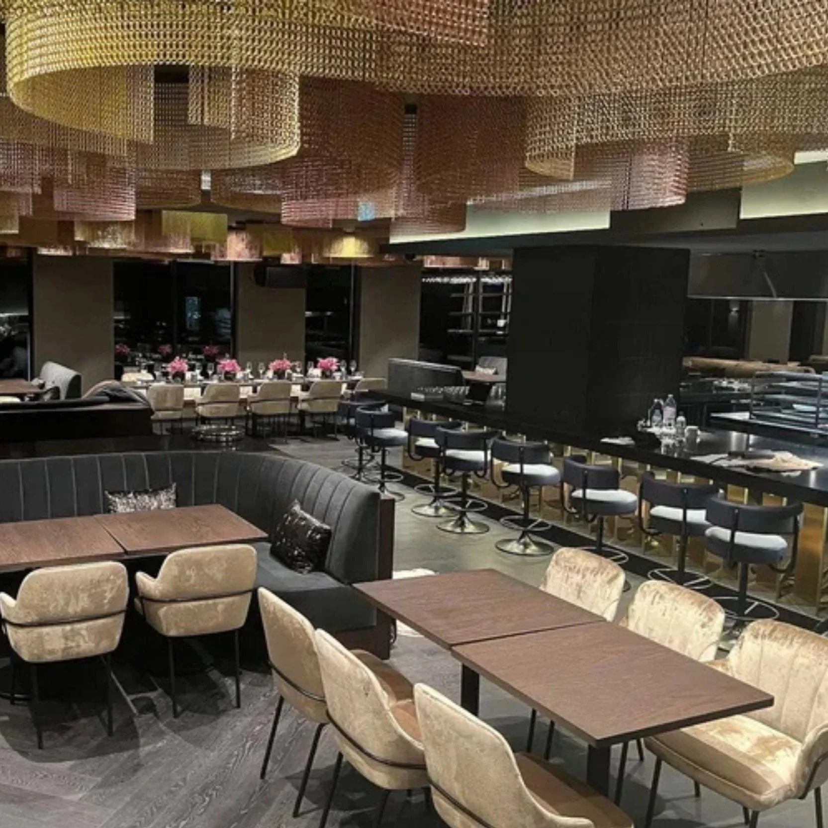 Elegant restaurant interior featuring beige upholstered chairs, a large meeting table, a curved booth with pillows, bar seating with black stools, floral arrangements, and a decorative ceiling with textured panels.
