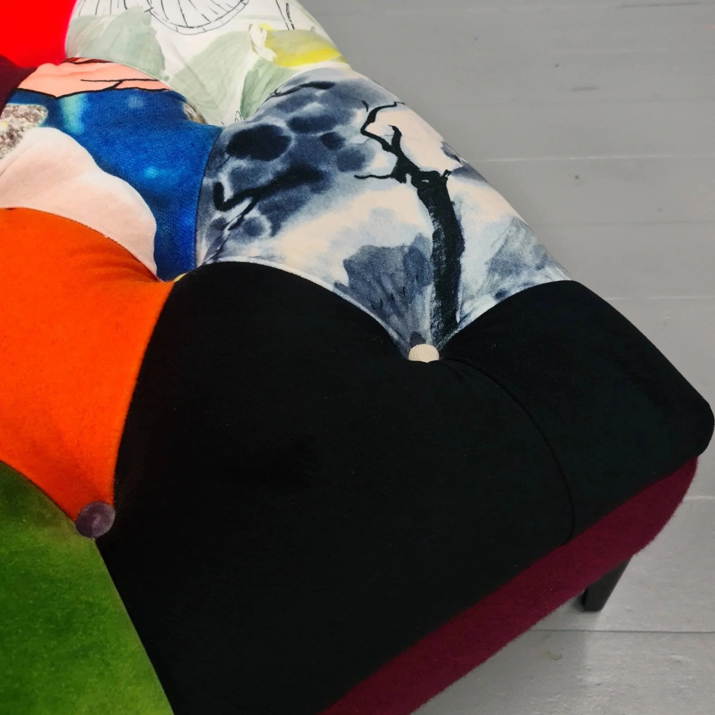 Close-up of a colorful patchwork velvet armchair with various patterned fabrics on the back and seat, set against a gray wall.