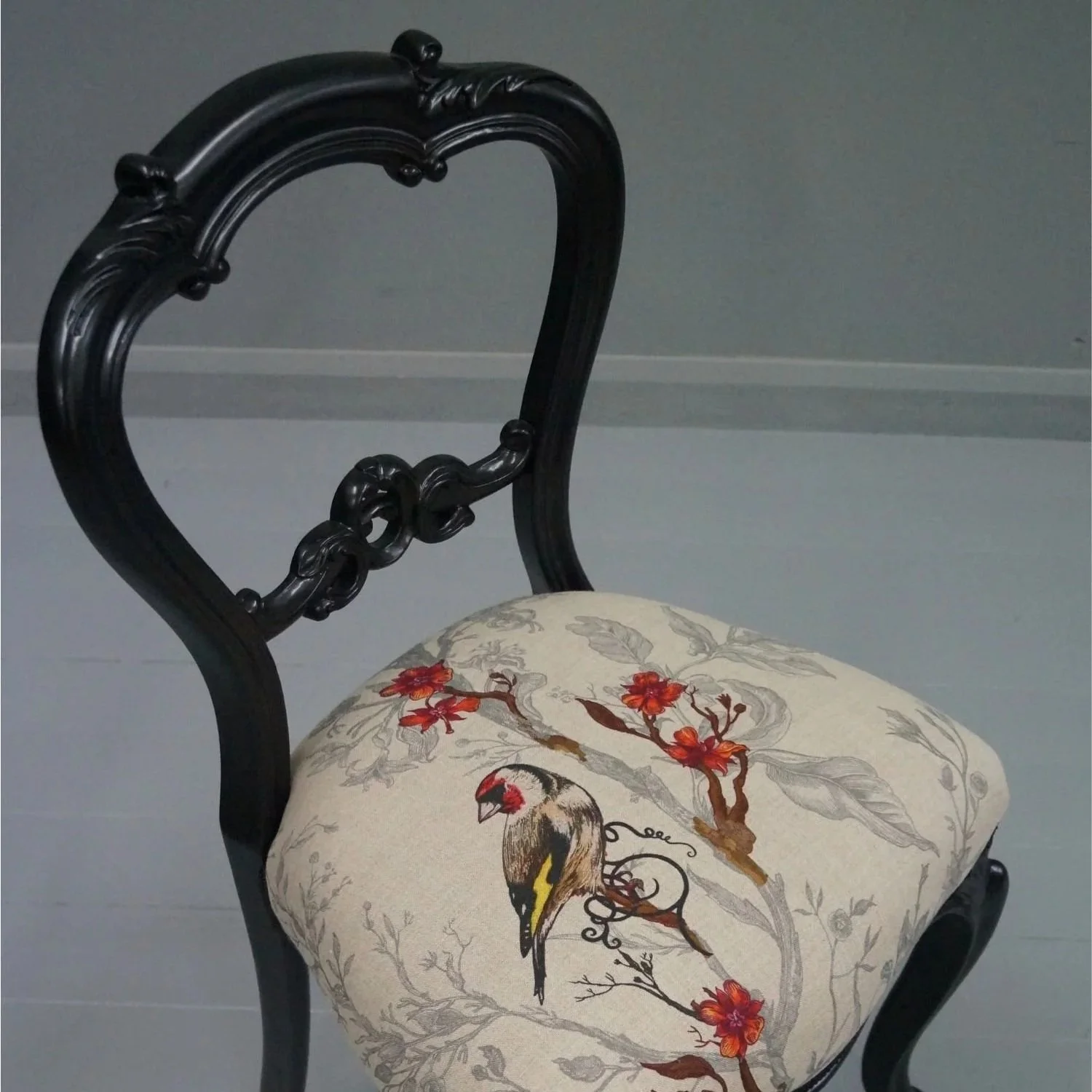A black wooden chair with ornate carved backrest, featuring a cushion with a bird, red flowers, and grey foliage pattern.