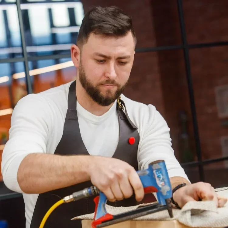 Man in a gray apron using a hot glue gun on a piece of wood in a workshop.