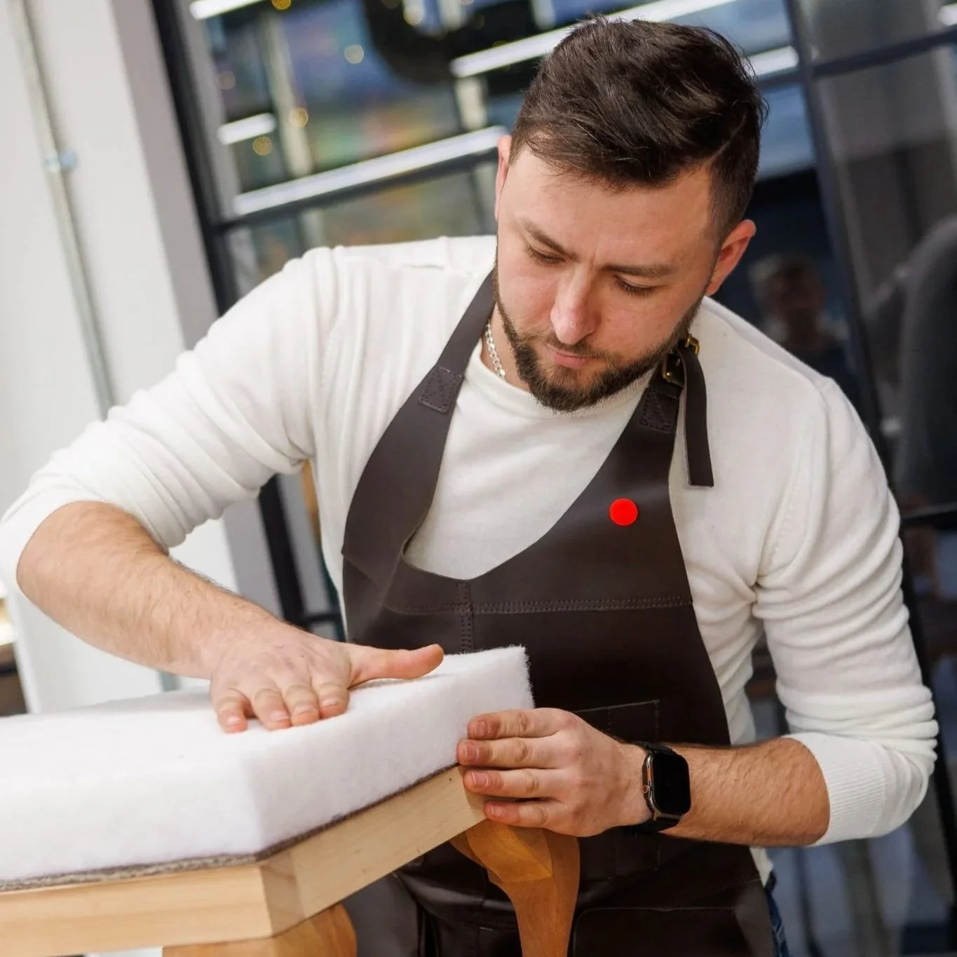A man with a beard and short hair, wearing a white long-sleeve shirt, a black apron, and a watch, is polishing a wooden table with a white cloth in a modern indoor setting.