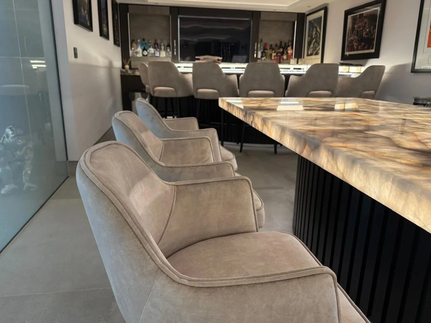 A modern home bar area with plush beige chairs lined up in front of a large marble countertop. The bar has a variety of liquor bottles and artwork on the walls.