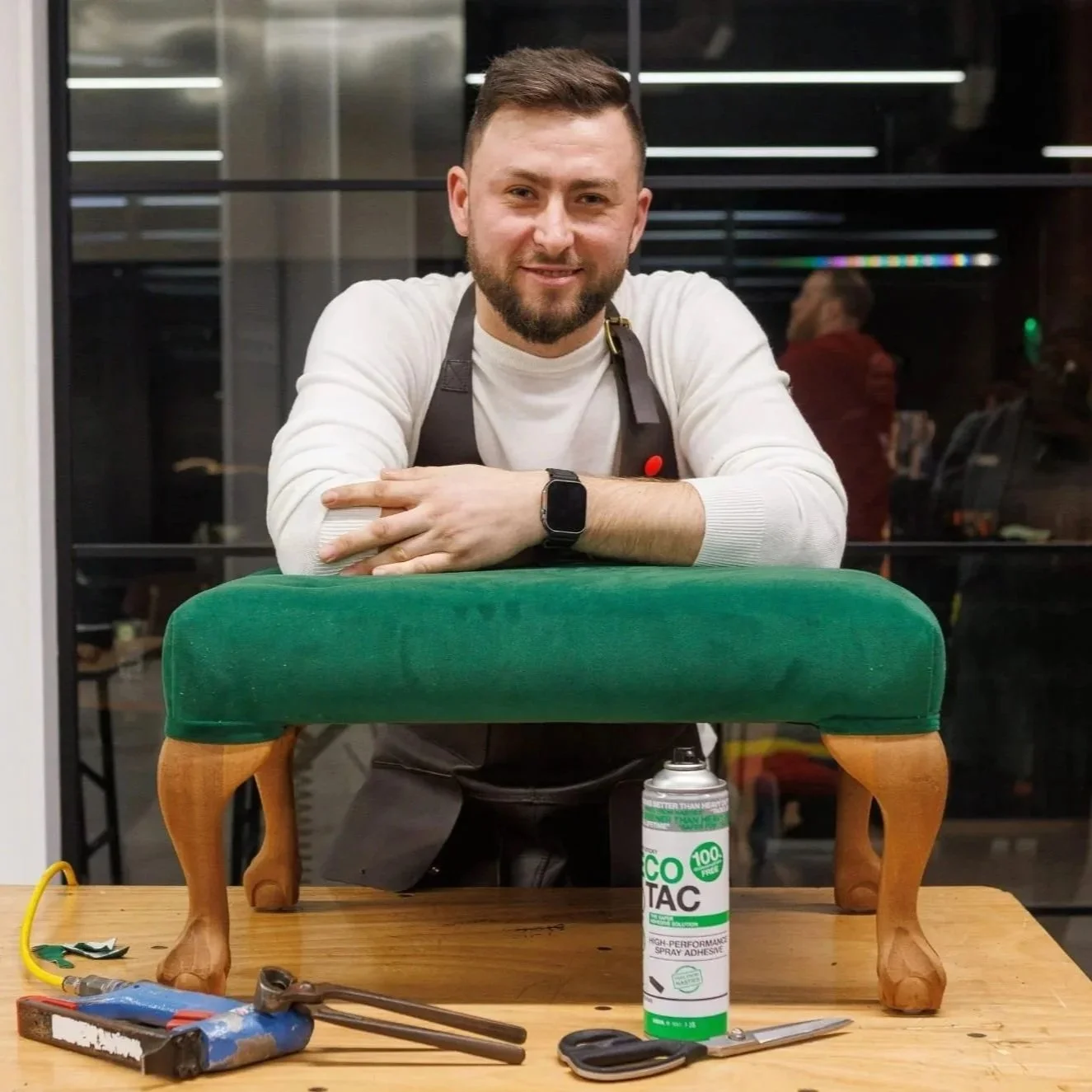 A man with a beard smiling and leaning on a green upholstered chair with wooden legs, on a wooden table with tools, burst spray adhesive, scissors, a wrench, and wires, in a workshop or studio.