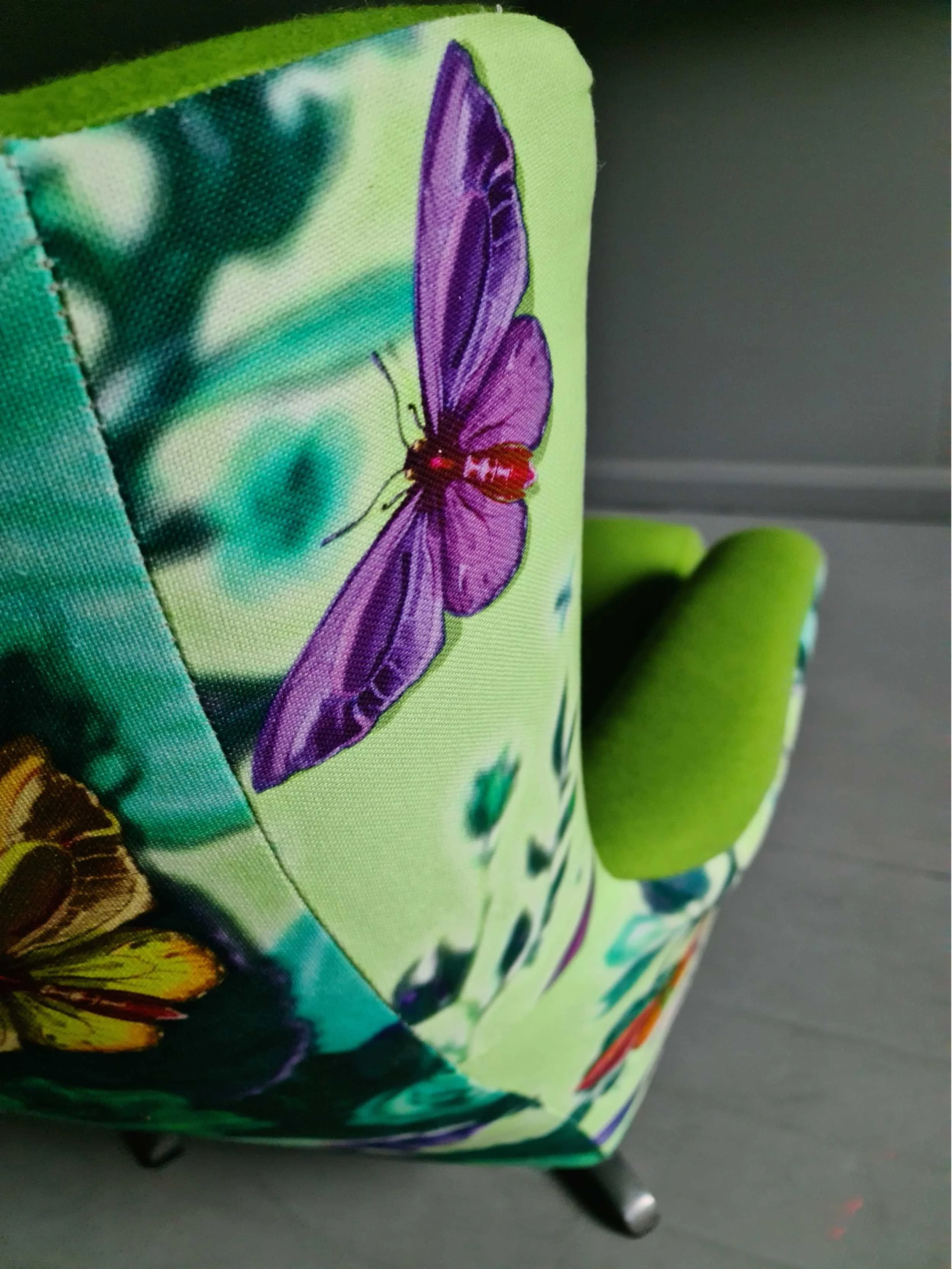 Close-up of a upholstered chair featuring a colorful butterfly print with purple, yellow, and orange butterflies against a vibrant green and teal background.