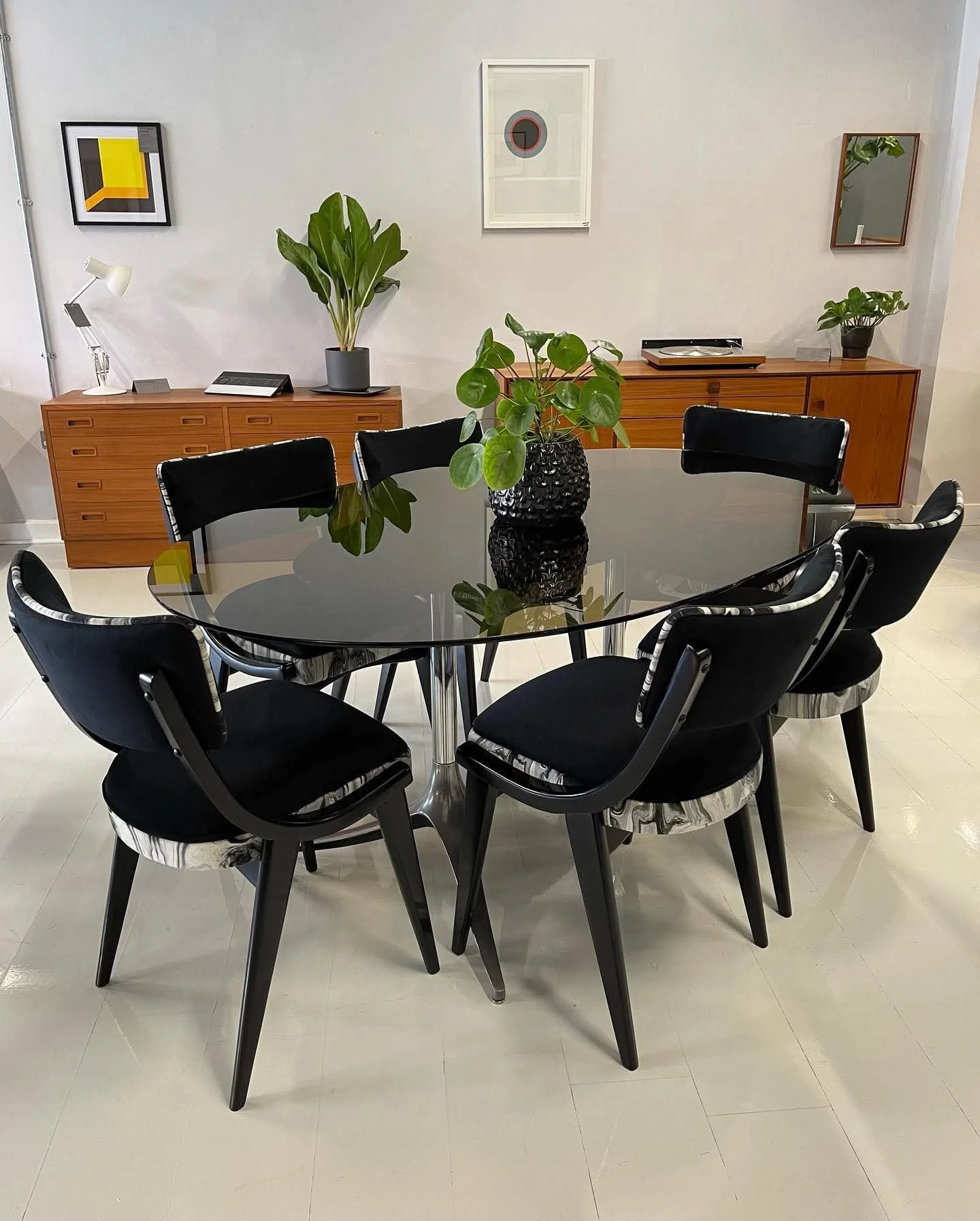 A modern dining room with a black glass dining table, six black chairs with white and black marble design accents, and a black textured vase with a green plant on the table. In the background are two wooden sideboards with plants, decorative items, and artwork on the wall.