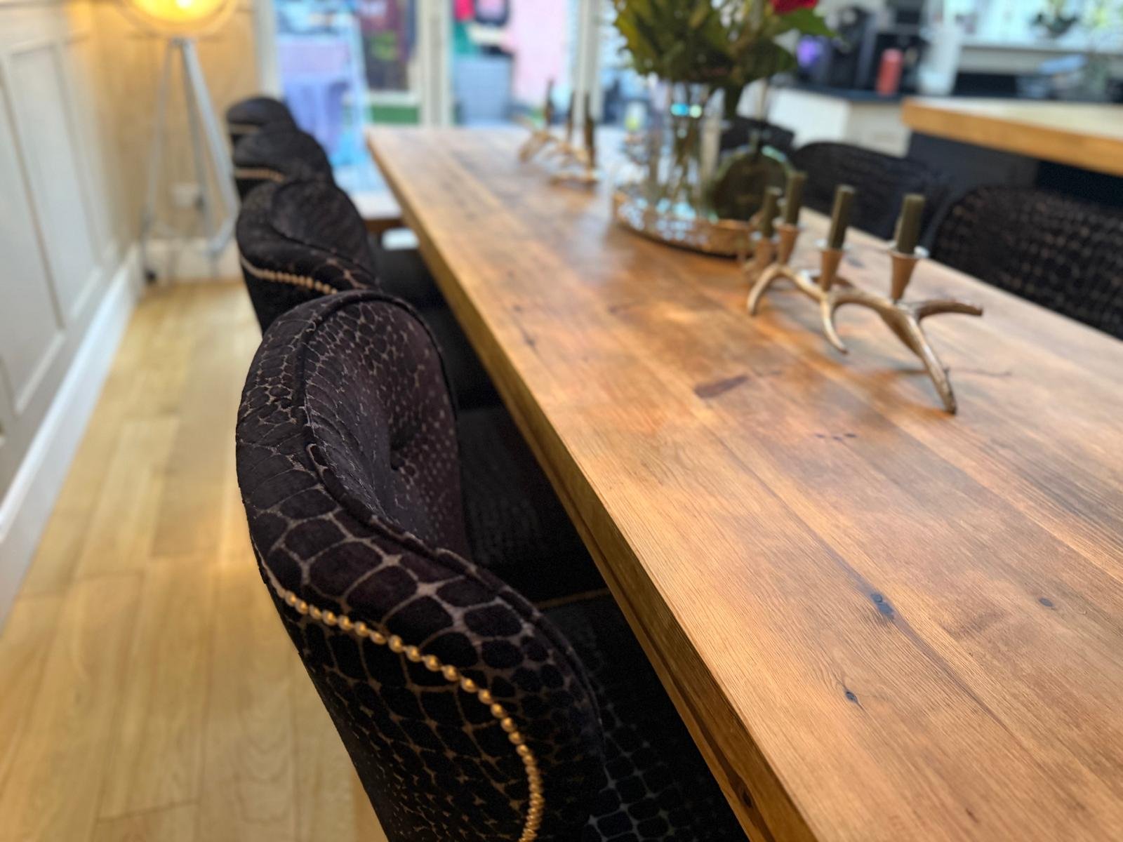 A wooden dining table with black upholstered chairs with gold nailhead trim, decorated with a antler-shaped candelabra and a centerpiece, in a bright room with hardwood floors and large windows.