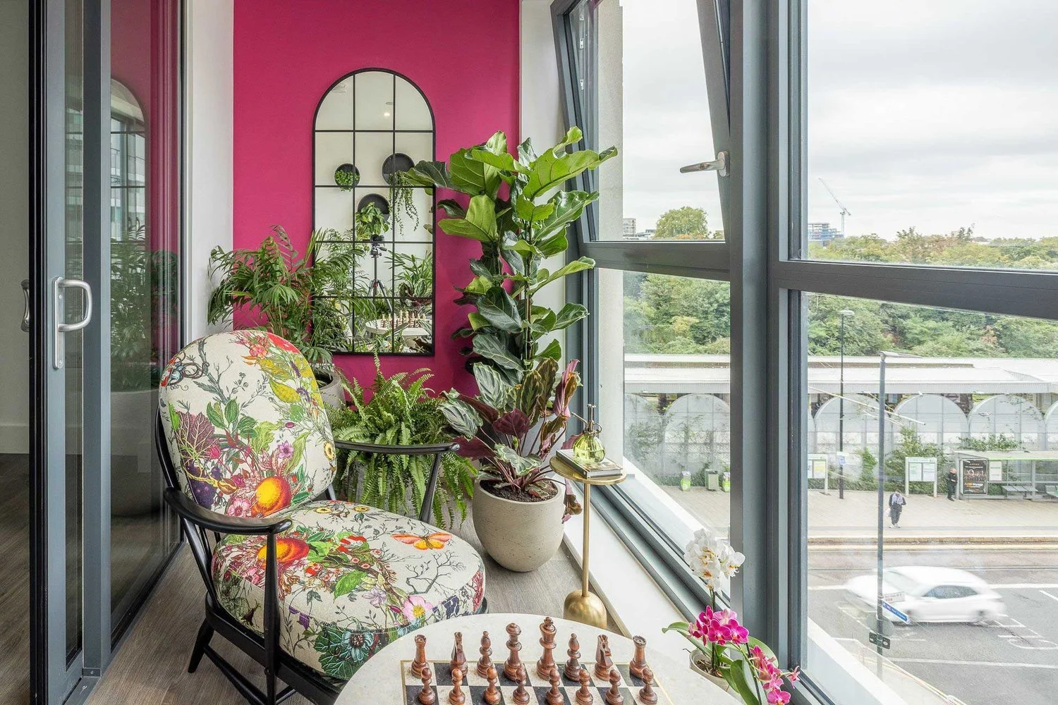 Sunlit balcony with a colorful floral upholstered chair, large potted plants, a gold floor lamp, a small side table with a glass bottle, a chessboard, and panoramic city view through glass windows.