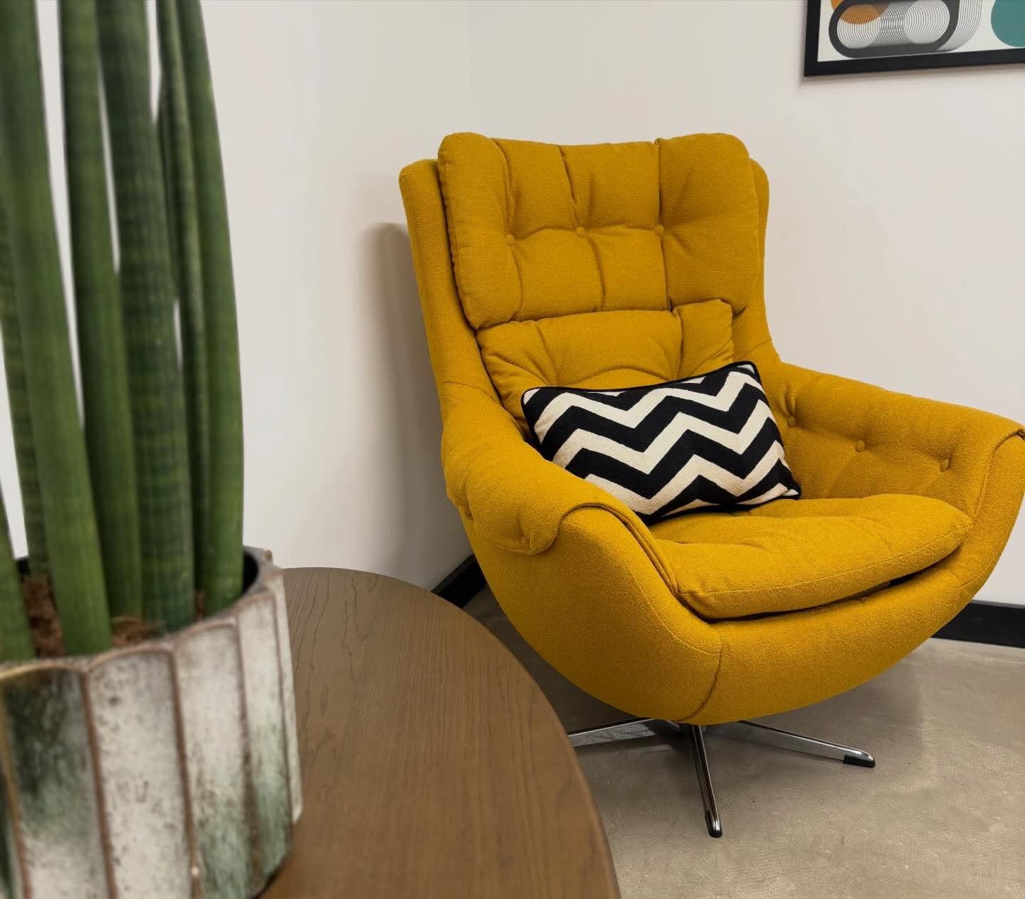 A yellow mid-century modern wingback chair with a black and white chevron pillow in a room with a potted plant on a wooden table and a white wall.