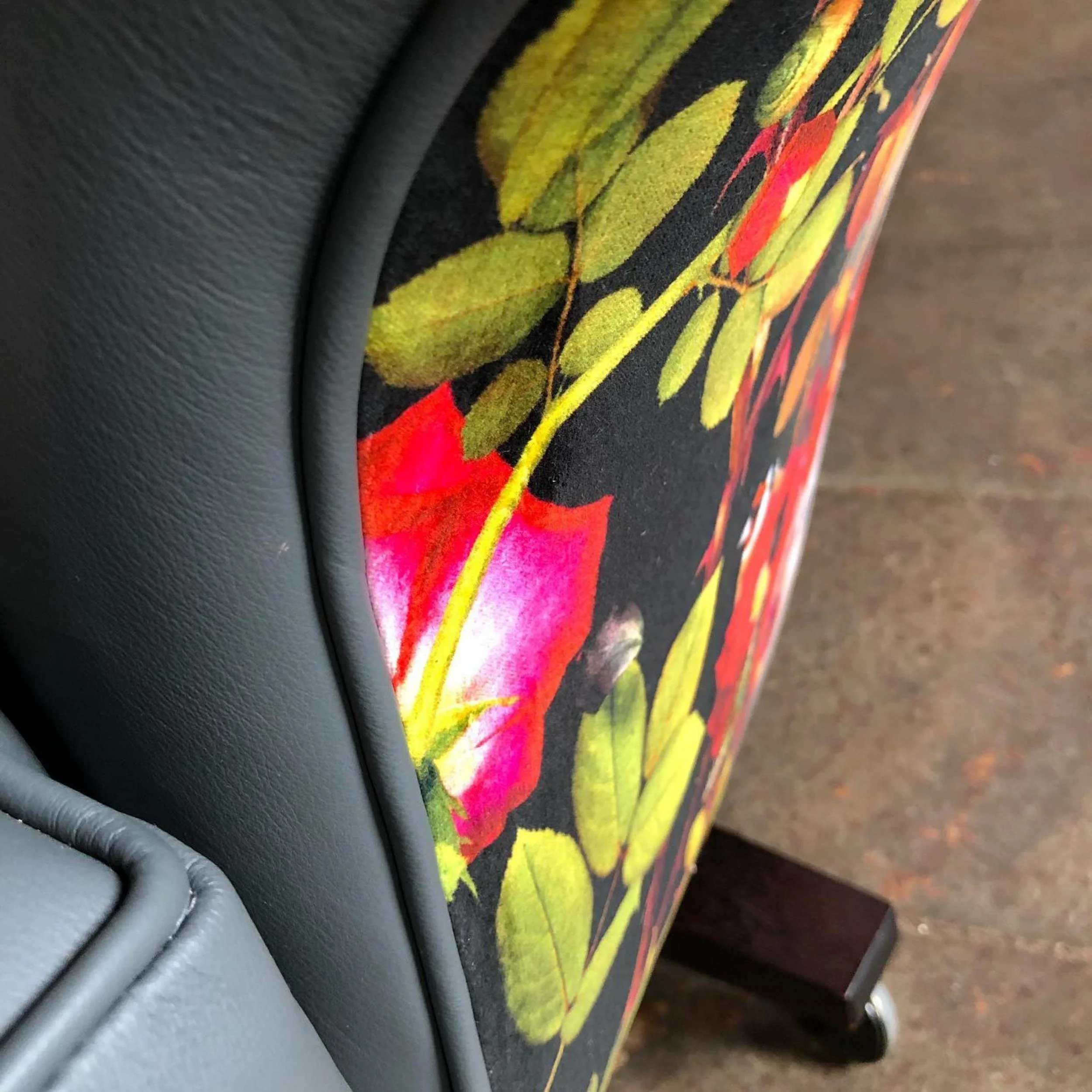 Close-up of a piece of furniture with a floral fabric featuring pink, orange, and yellow flowers with green leaves, and a black background. The image is taken at an angle showing the side and part of the back of the furniture, which has leather or faux leather trim.