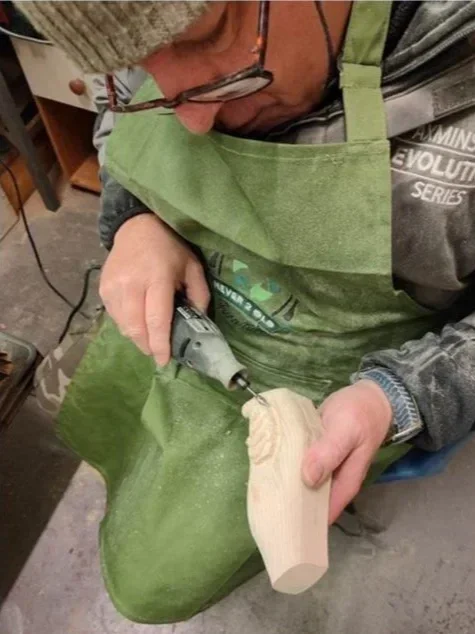Person carving into a piece of light-colored wood using a rotary tool, wearing glasses, a green apron, and a gray sweater.
