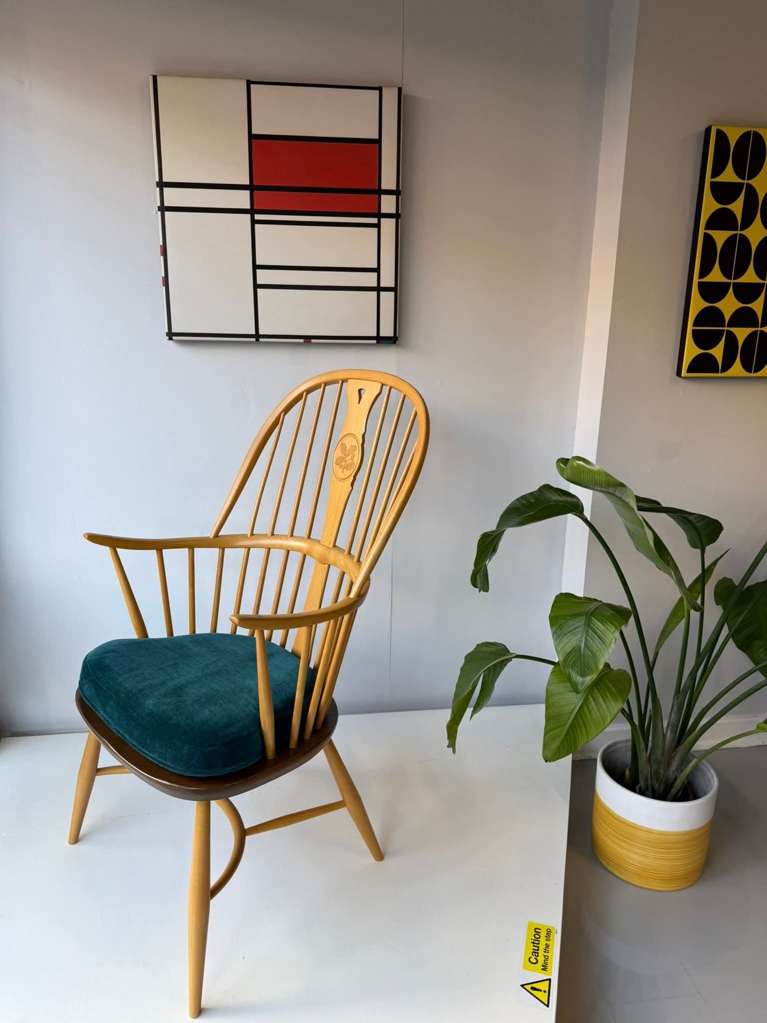 A wooden chair with a teal cushion next to a large green potted plant, on a white floor, with abstract art on the wall, in an indoor space.