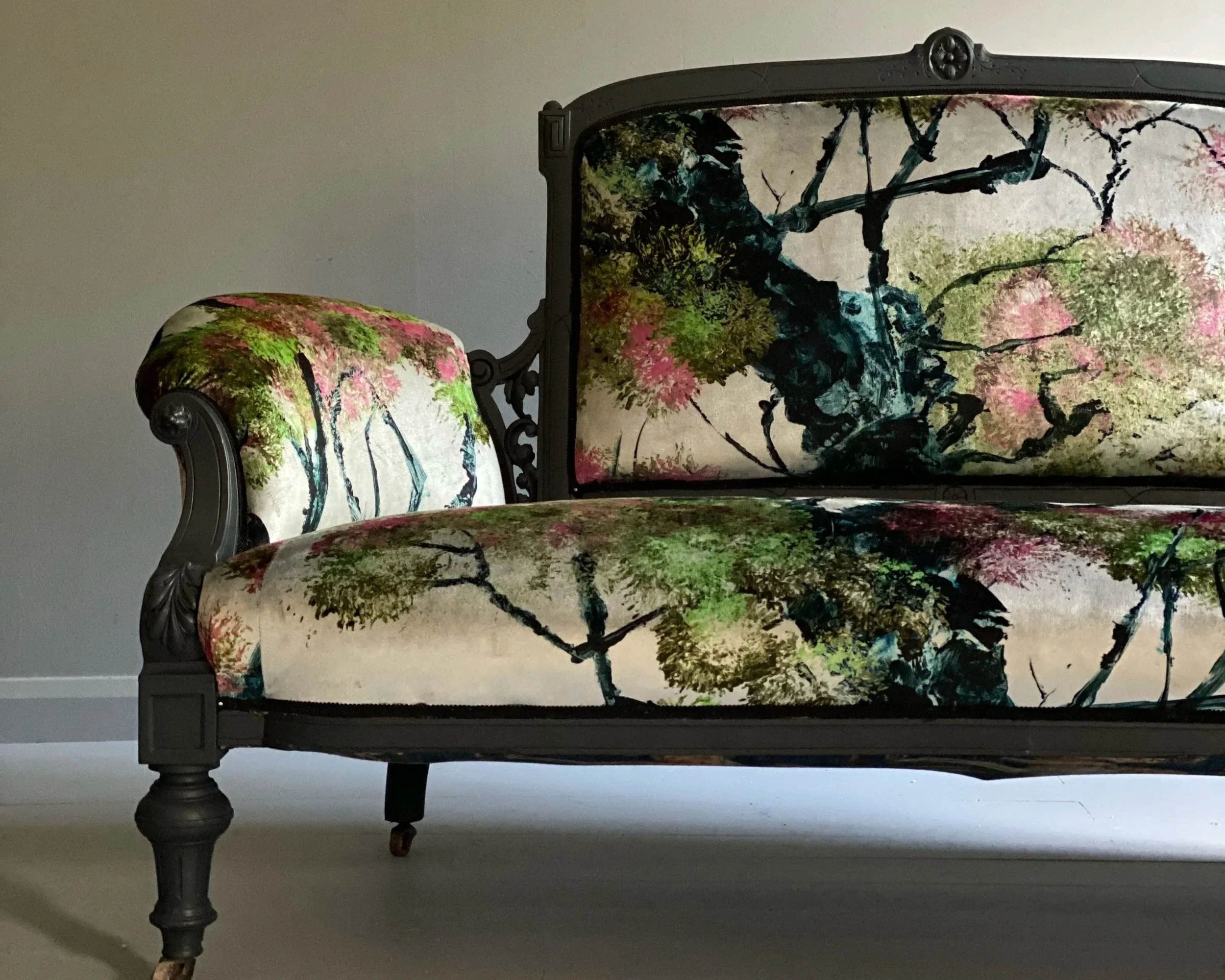 An ornate vintage sofa and matching armchair with wooden frames and legs, upholstered in fabric with a floral pattern featuring trees and branches in pink, green, and black colors.