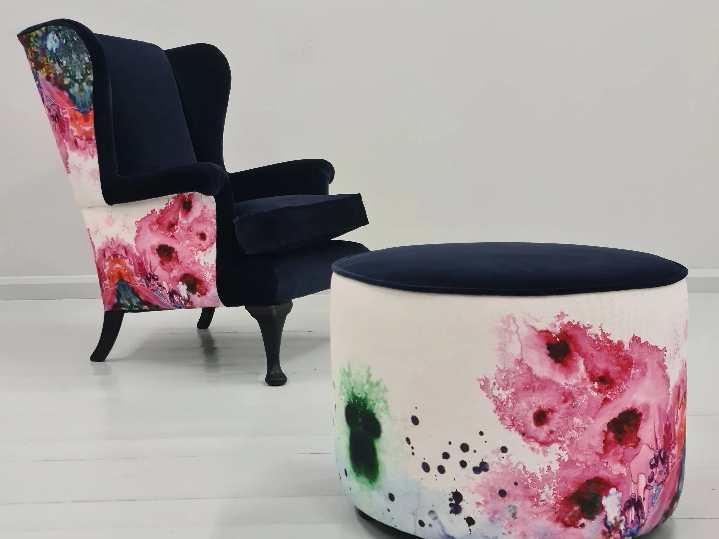 A dark blue armchair with floral fabric on the sides and a matching round ottoman, both featuring pink floral watercolor patterns, set against a white wall and floor.