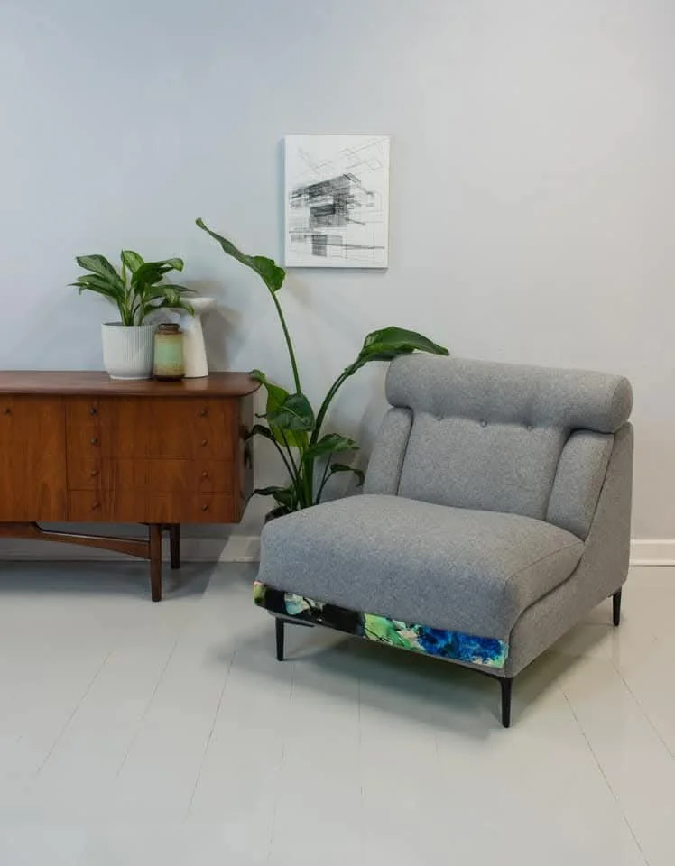 A cozy corner with a gray upholstered armchair, a wooden sideboard with potted plants and decorative vases, and abstract artwork on a white wall.