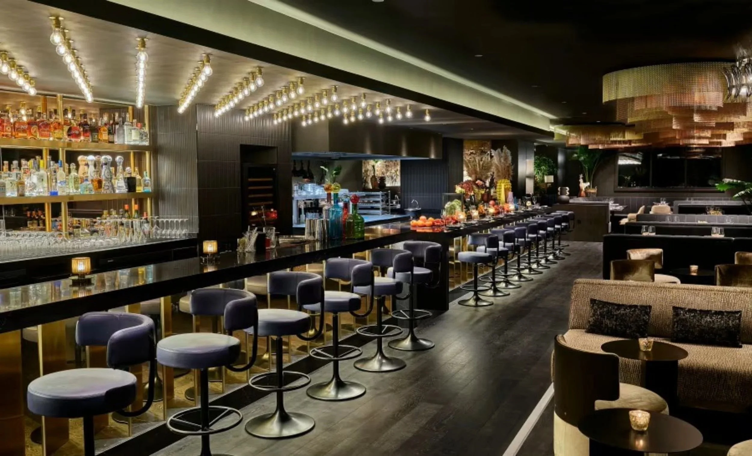 Elegant bar with black countertops, purple barstools, and shelves stocked with various liquor bottles, featuring modern lighting and a cozy seating area with candles.