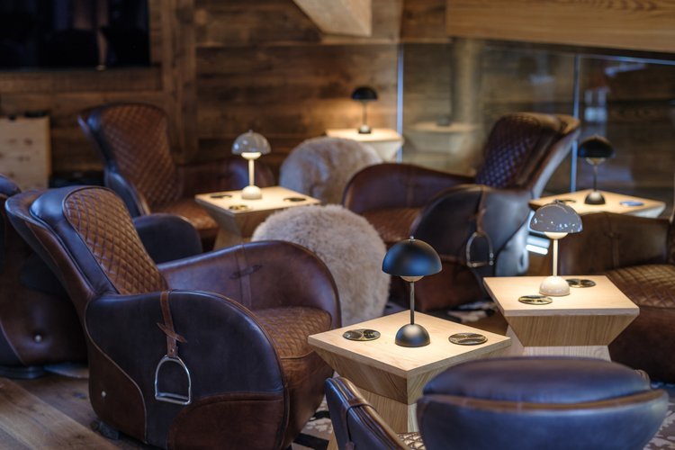 Cozy lounge area with leather armchairs, small wooden tables lit by desk lamps, in a rustic wooden interior.