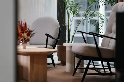 A modern indoor seating area with two beige armchairs, a wooden coffee table, a potted plant, and a large window with natural light.