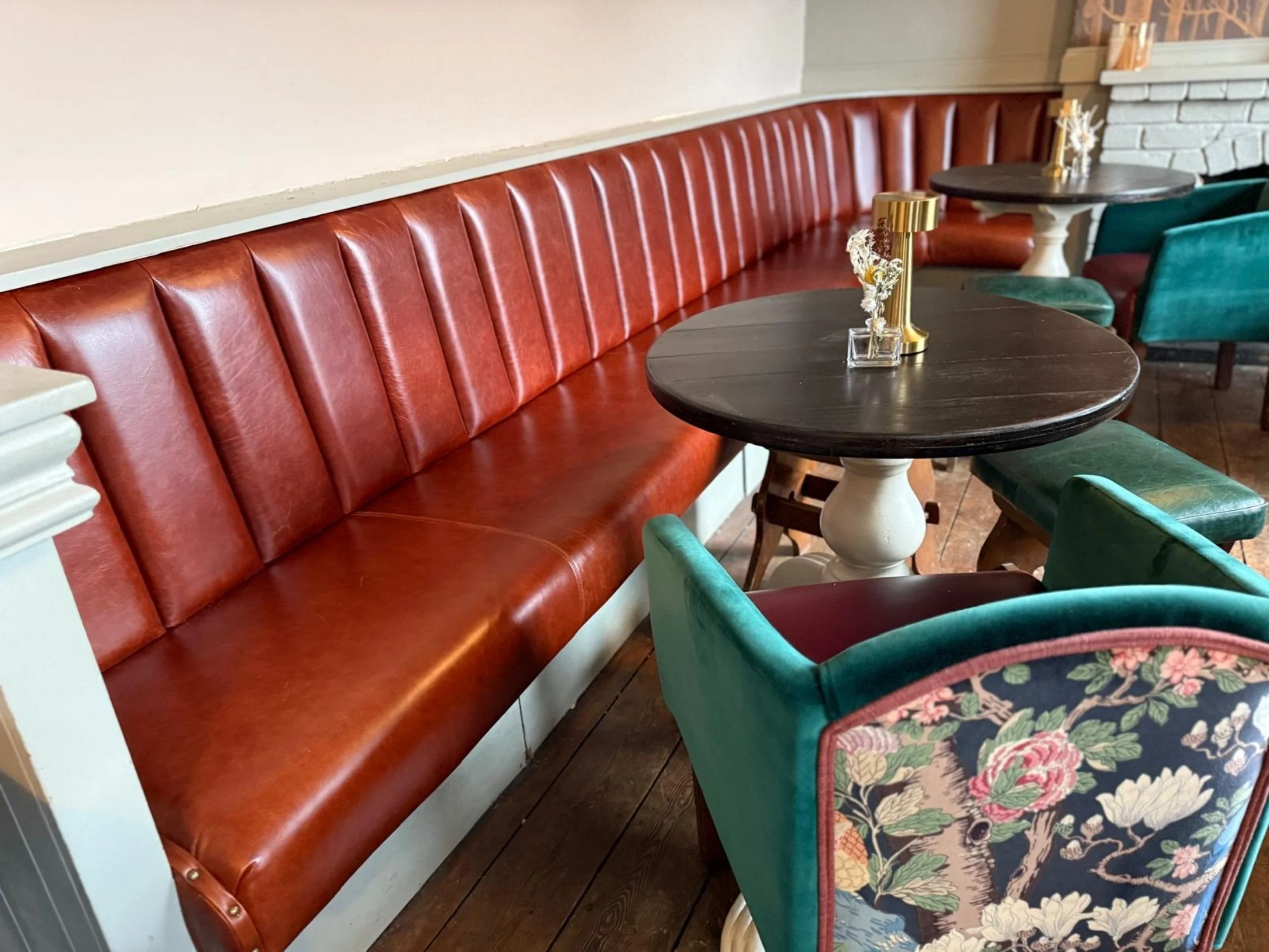 Restaurant interior with a long red leather bench seat, several round dark wood tables, and colorful upholstered chairs with floral and modern patterns.