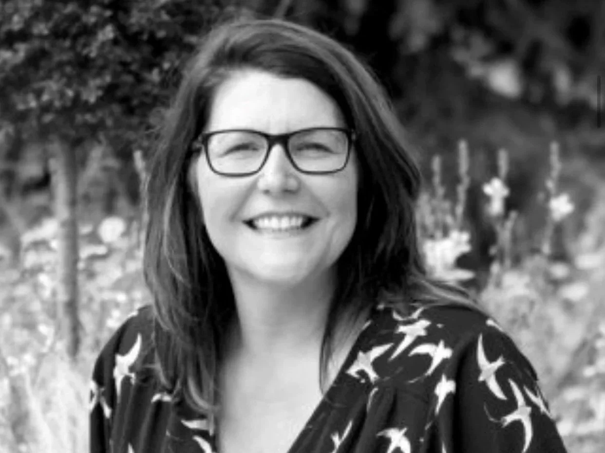 A woman with glasses and shoulder-length hair smiling outdoors, wearing a patterned dress with birds, with trees and plants in the background.
