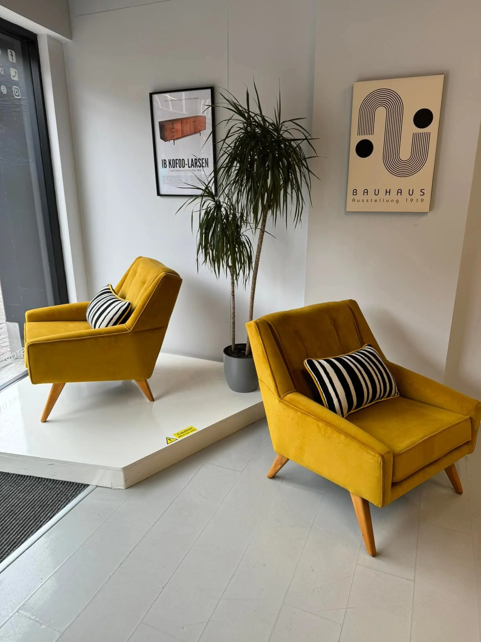 Two yellow armchairs with black and white striped pillows, a potted plant, and framed posters on the wall inside a modern seating area.