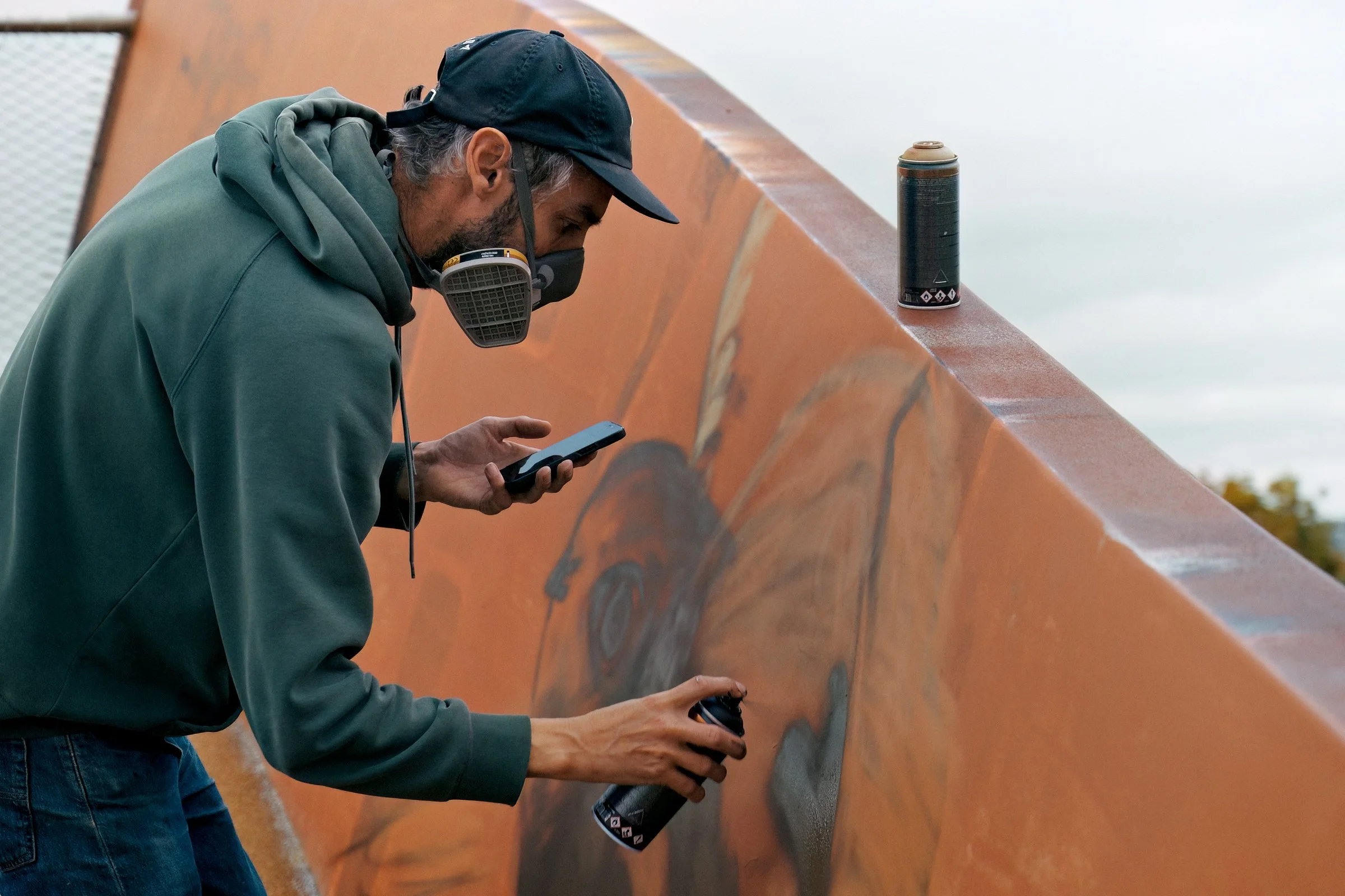 Man wearing a green hoodie and black cap, with a dust mask, spray painting a mural on a large orange wall, using a black spray paint can, while looking at his phone.