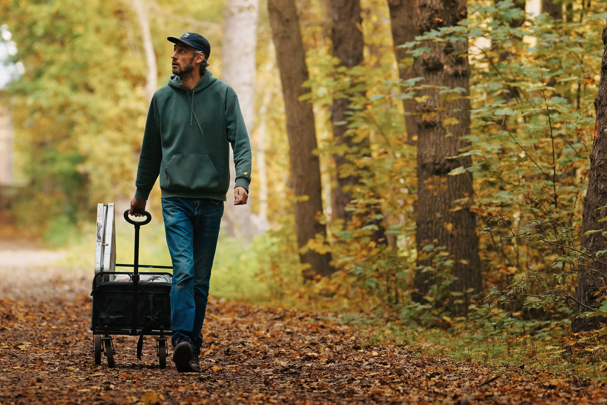 A man walking on a wooded trail during autumn, pulling a black rolling suitcase with a backpack attached, surrounded by fall foliage.