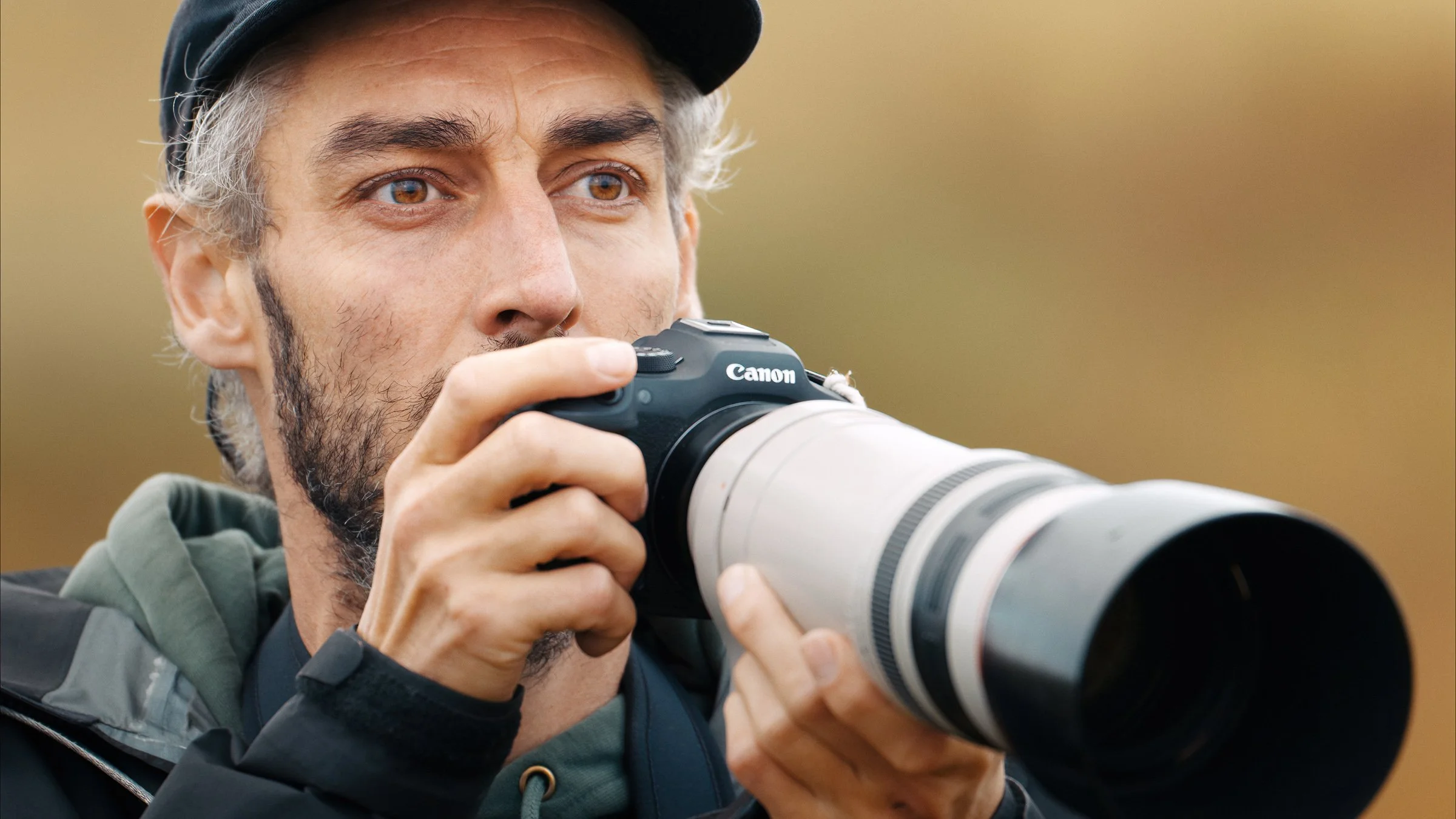 A man with a beard holding a Canon camera with a large telephoto lens, looking through the camera's viewfinder outdoors.