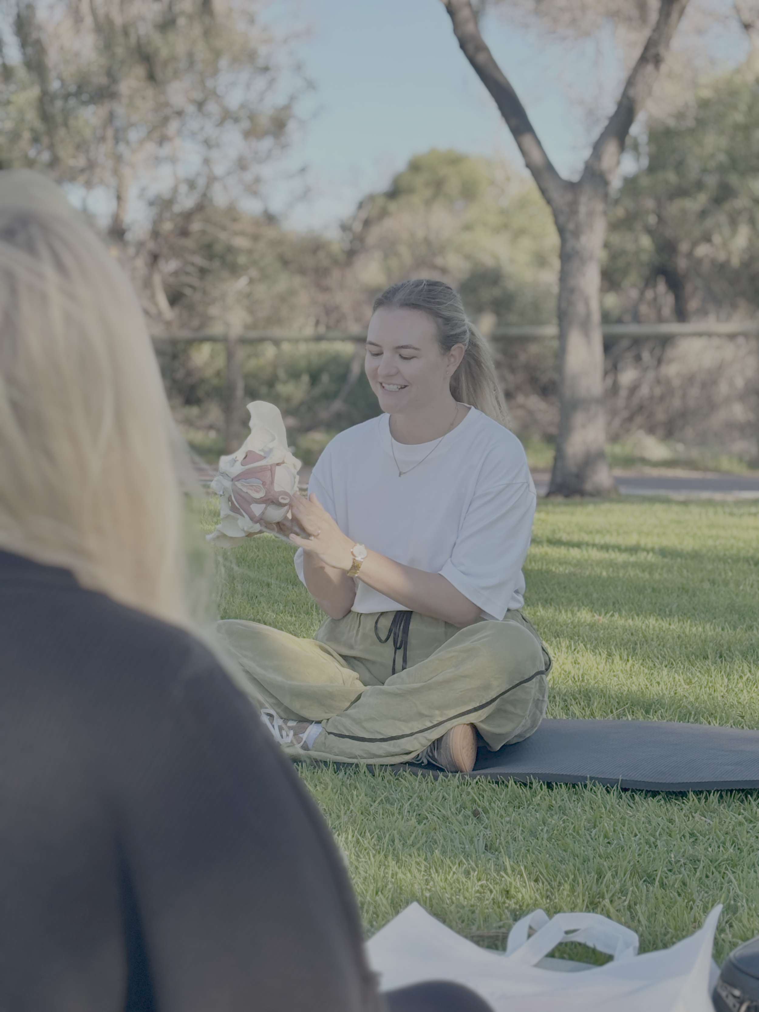 Young woman sitting on the grass outdoors, holding a model of a human heart, smiling, with trees in the background.