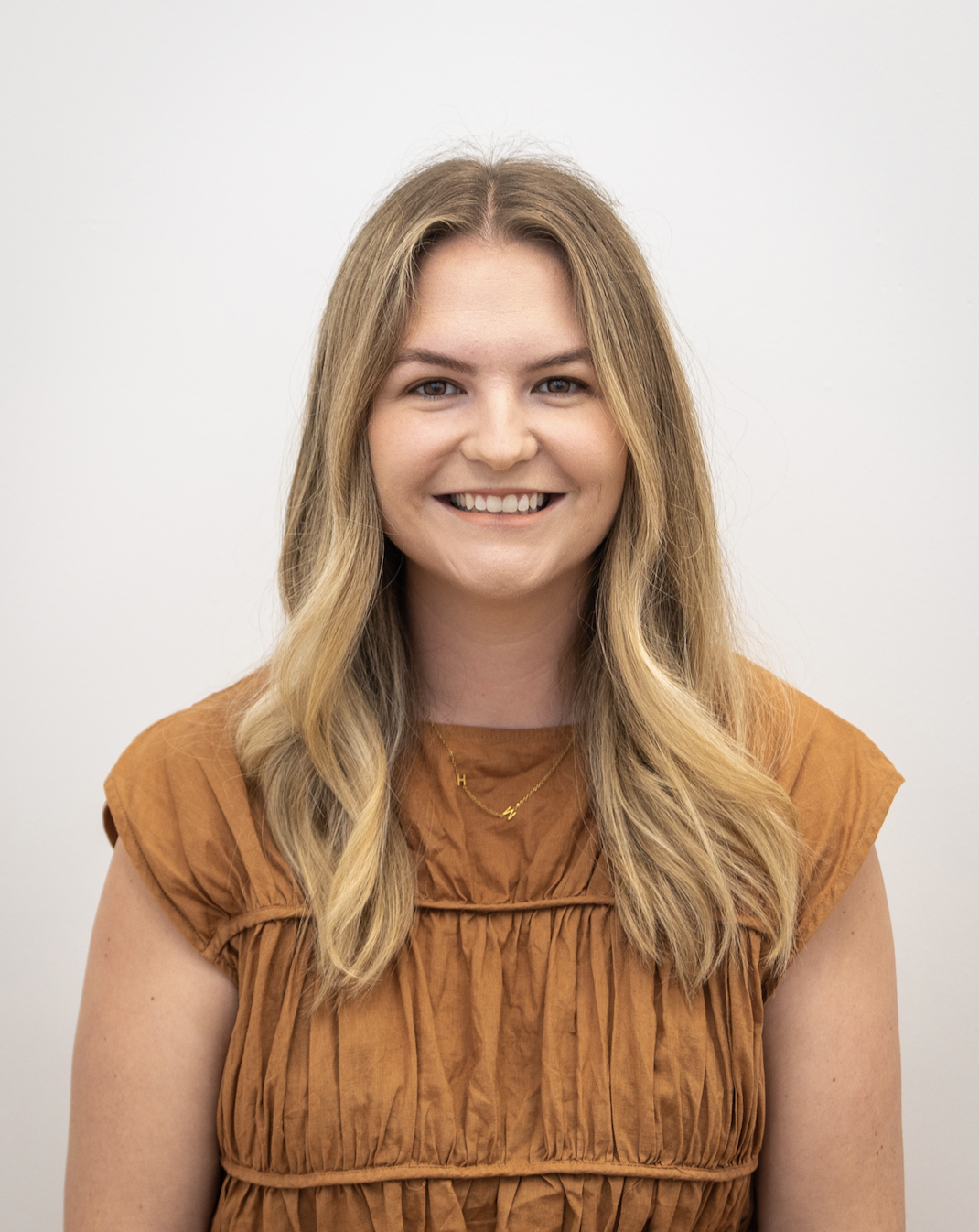 Close-up of a young woman with long, wavy blonde hair smiling against a plain white background, wearing an orange top and gold necklaces.
