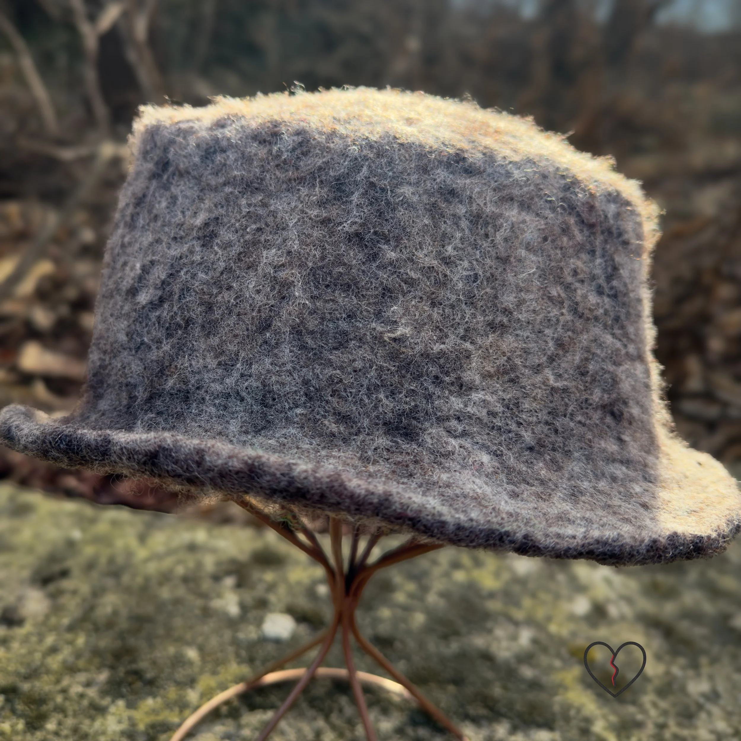 Hand-felted wool fedora hat in natural brown tones with a white and dark brown spiral design, sculptural crown, and short brim, photographed outdoors.