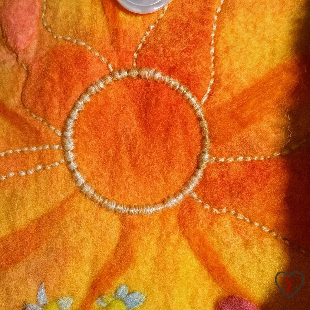 Close-up of embroidered circular sun motif on a hand felted wool bag, with stitched rays radiating across hand-dyed orange and yellow merino wool.