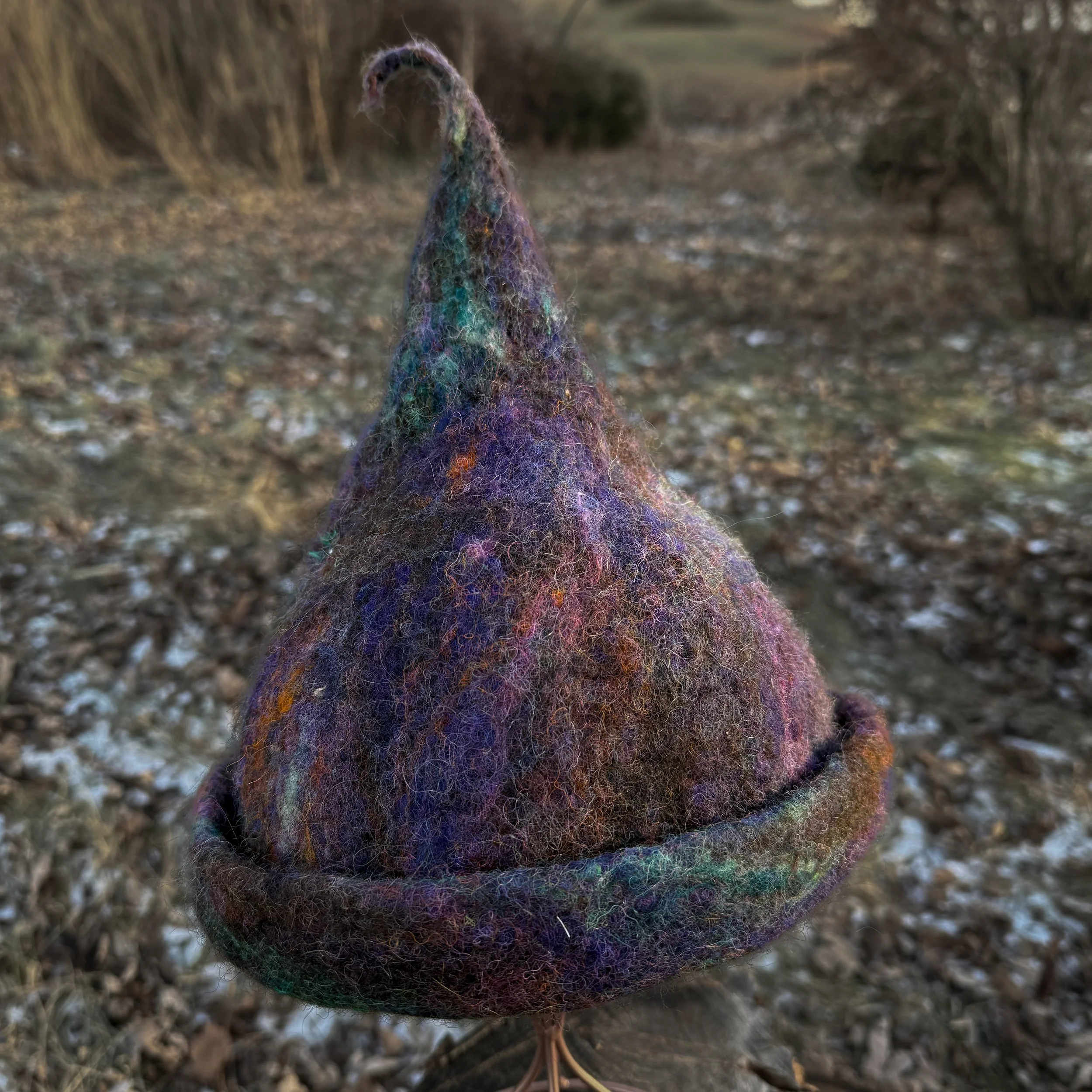 Purple and black hand-felted trainee witch hat with a curved tip and rolled brim, displayed on a wire stand outdoors.