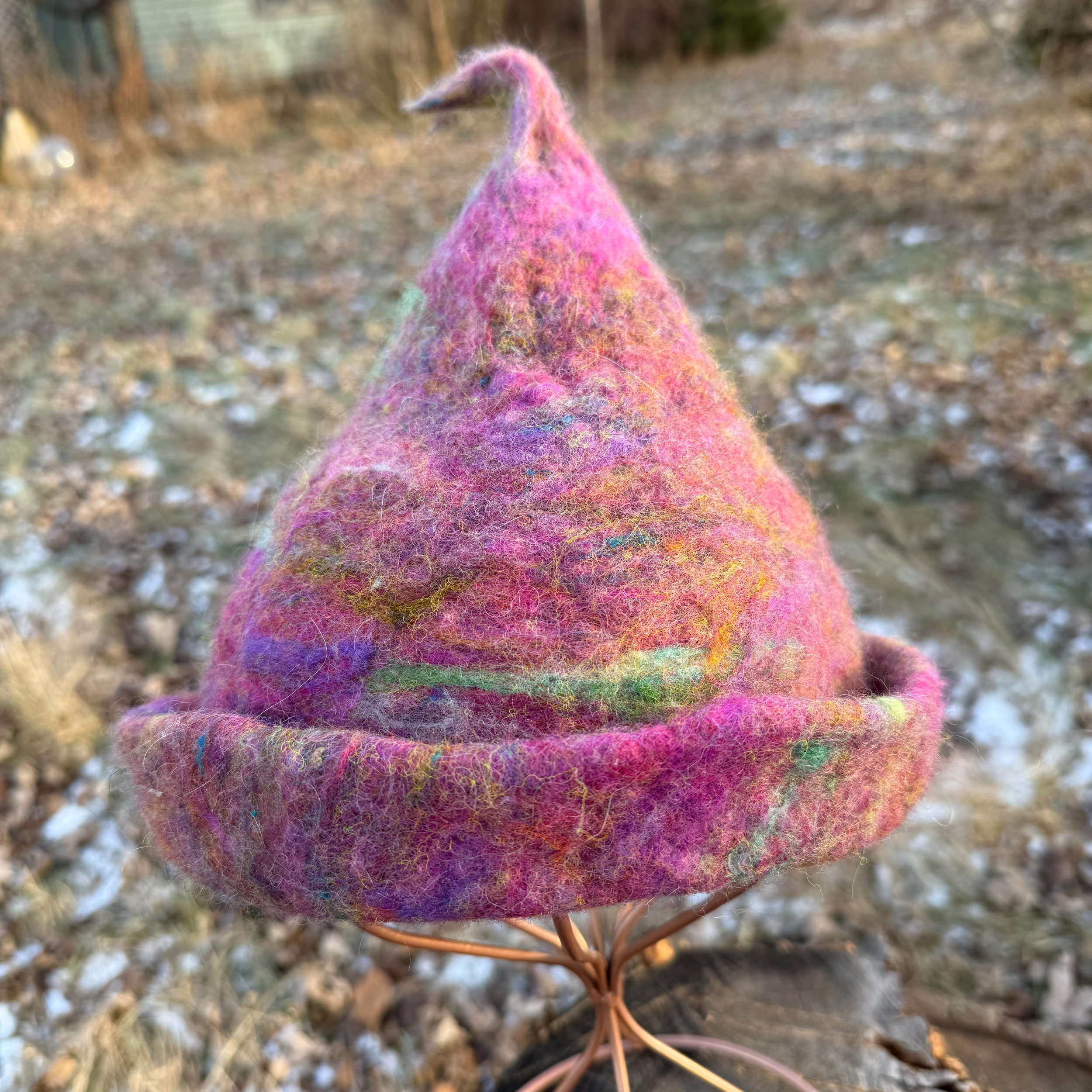 Pink hand-felted trainee witch hat with a curved tip and rolled brim, displayed on a wire stand outdoors.