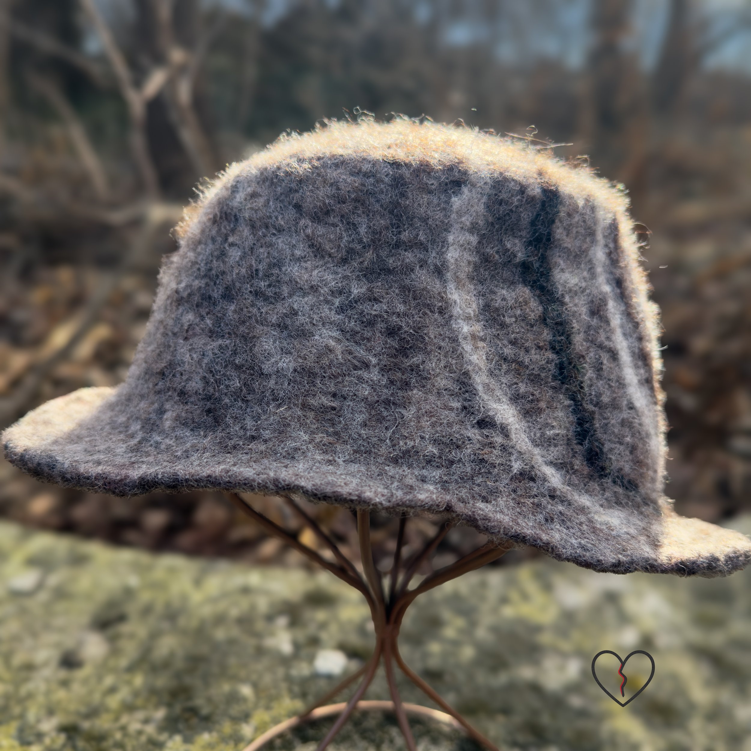 Hand-felted wool fedora hat in natural brown tones with a white and dark brown spiral design, sculptural crown, and short brim, photographed outdoors.