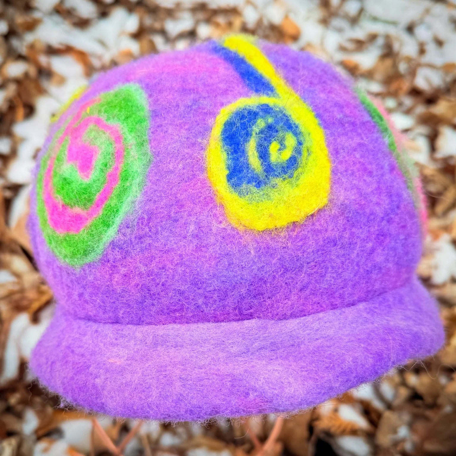 Handmade purple felted wool hat with small rolled brim and spiral designs, displayed on a stand outdoors in winter
