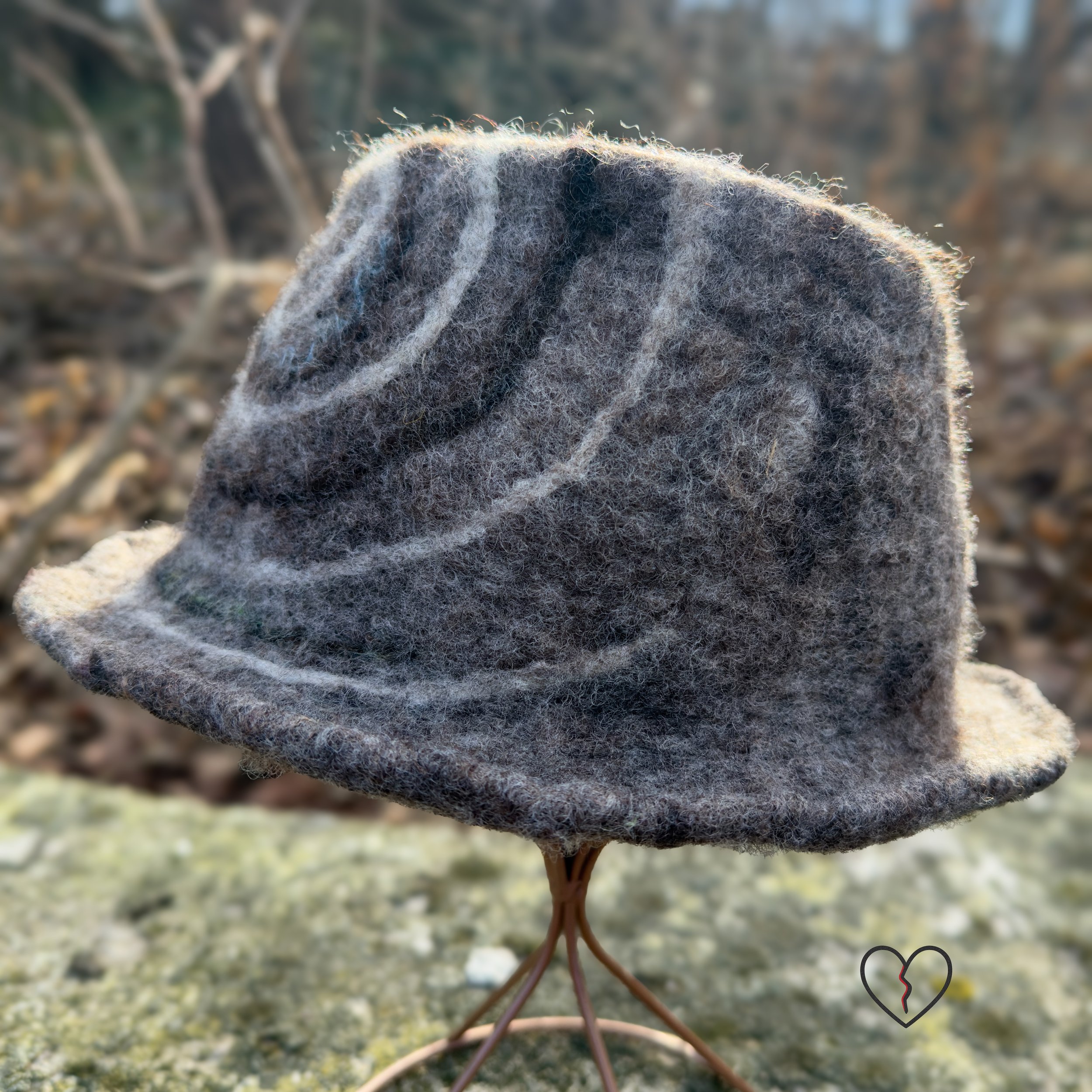 Hand-felted wool fedora hat in natural brown tones with a white and dark brown spiral design, sculptural crown, and short brim, photographed outdoors.