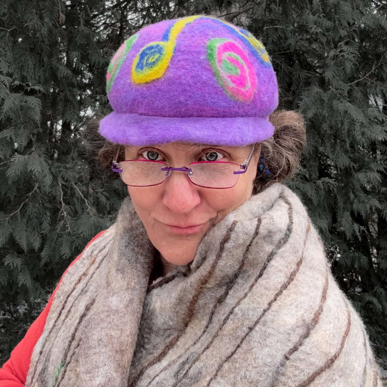 Fiber artist wearing a purple felted wool hat with triskele swirl design and rolled brim, photographed outdoors against evergreen trees