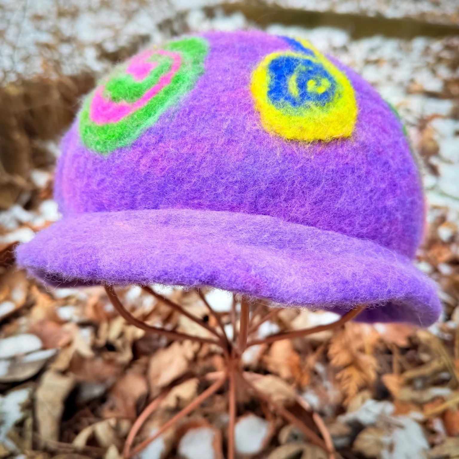Handmade purple felted wool hat with small rolled brim and spiral designs, displayed on a stand outdoors in winter