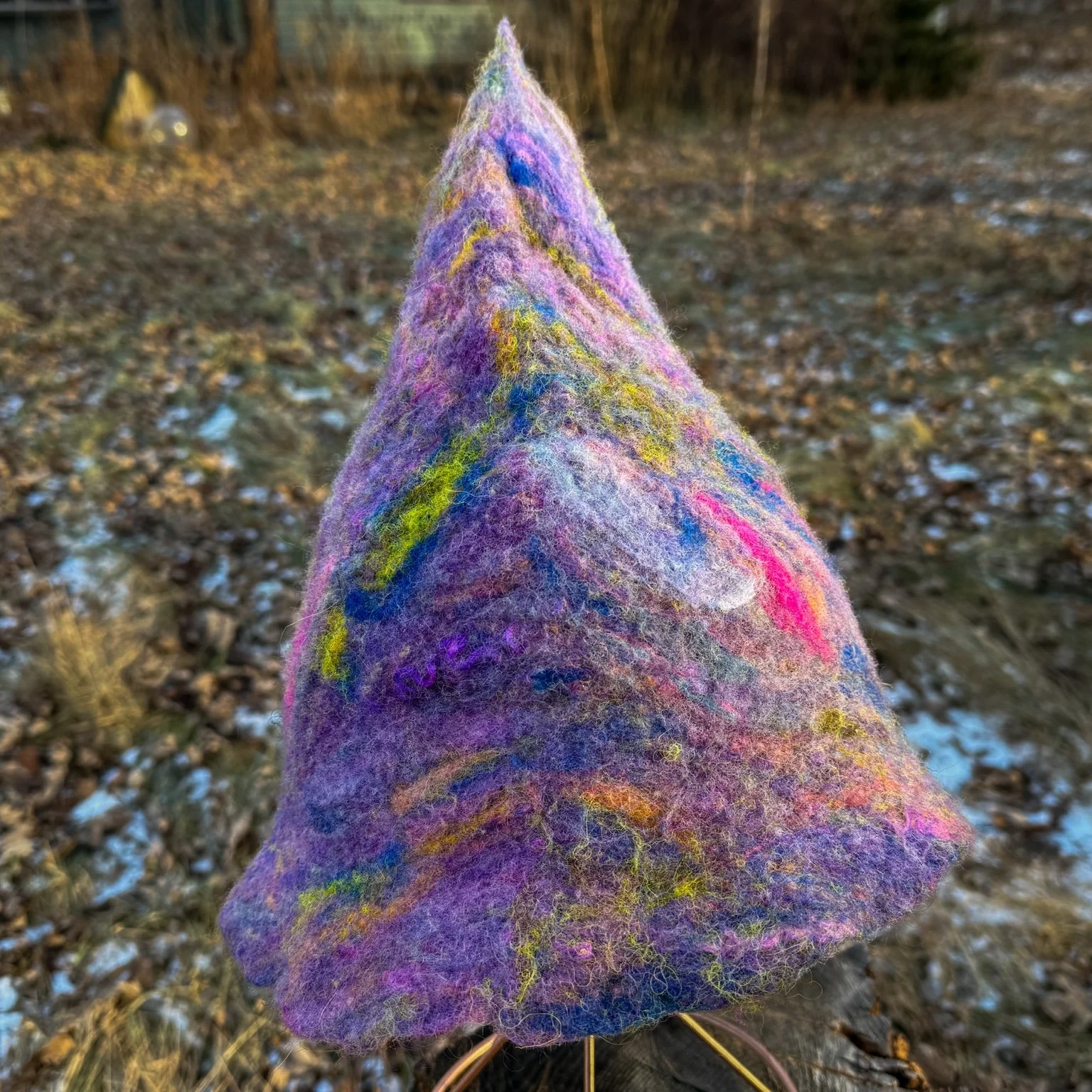 Hand-felted purple trainee witch hat with a tall pointed crown and natural edge, displayed on a wire stand outdoors.