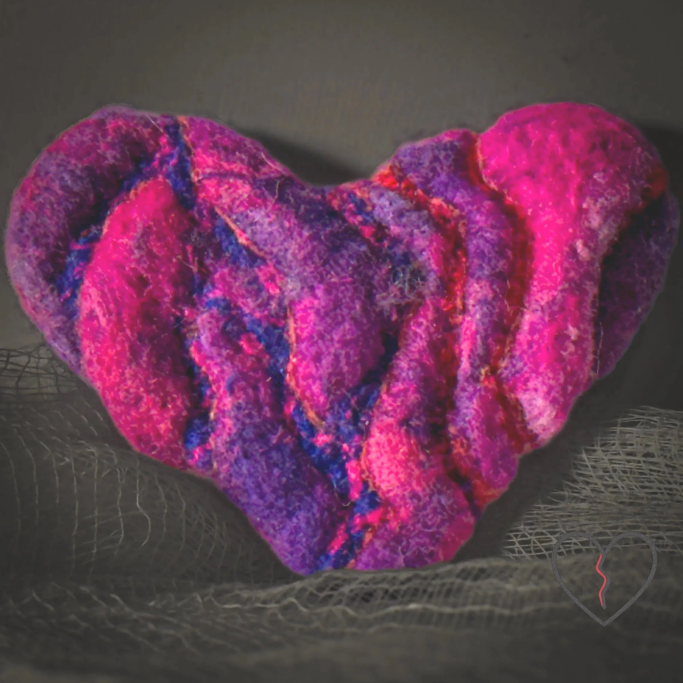 Hand-felted wool broken heart pin in pink and purple with blue and red lines running through textured cracks, photographed on dark fabric.
