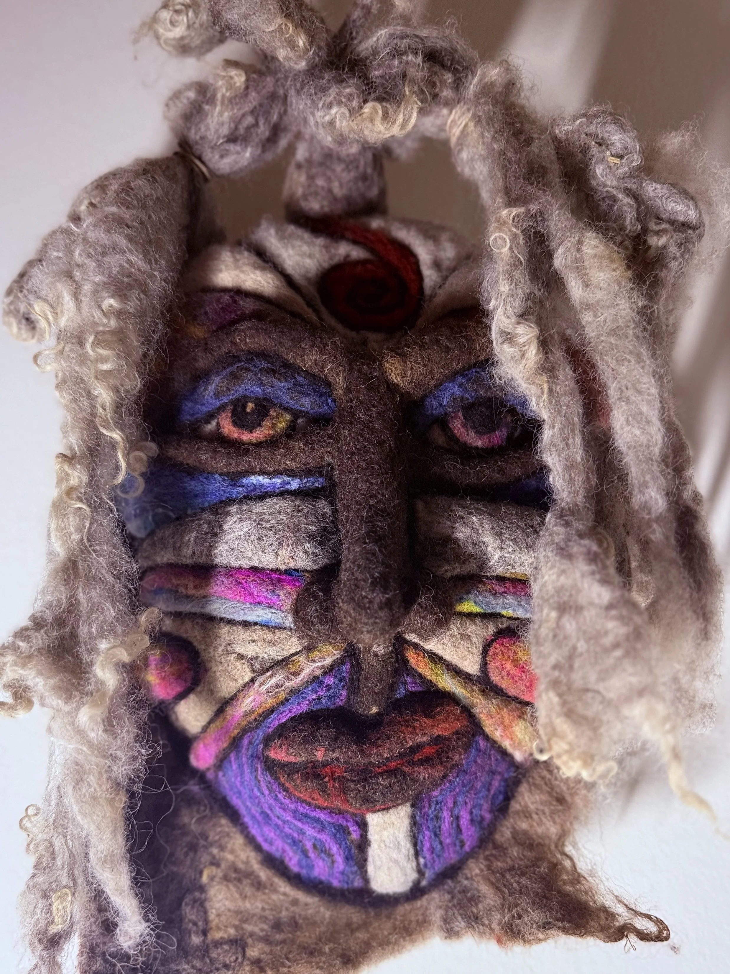 A handmade felted wool mask titled "Eira the Unyielding," featuring vivid sculpted details: a long central nose, deep-sett multicolored eyes, bold purple and pink markings, and textured grey wool hair cascading from the top.  The mask has a powerful,