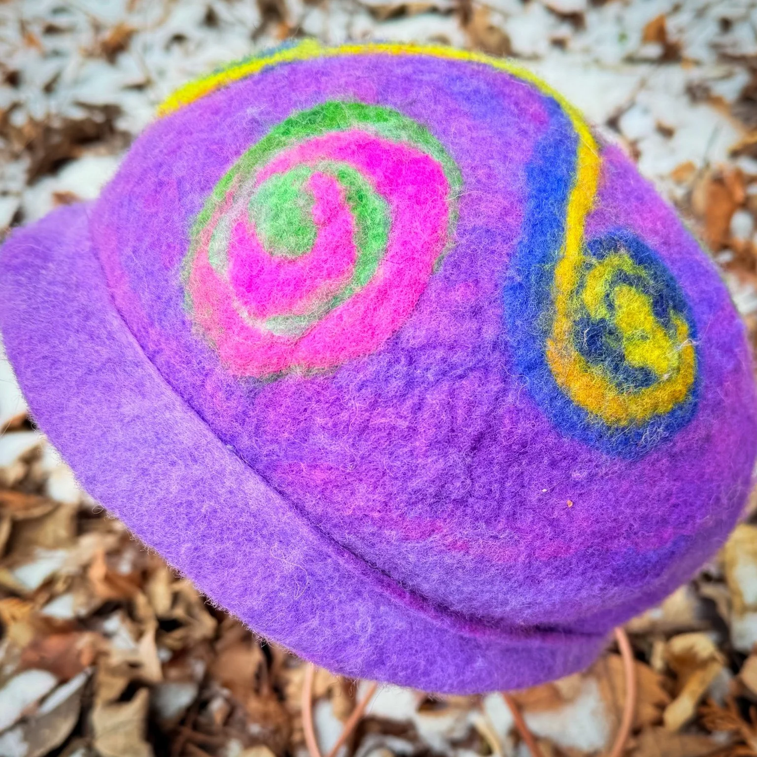 Angled top view of a one-of-a-kind felted wool hat featuring triskele-inspired movement and colorful spiral accents